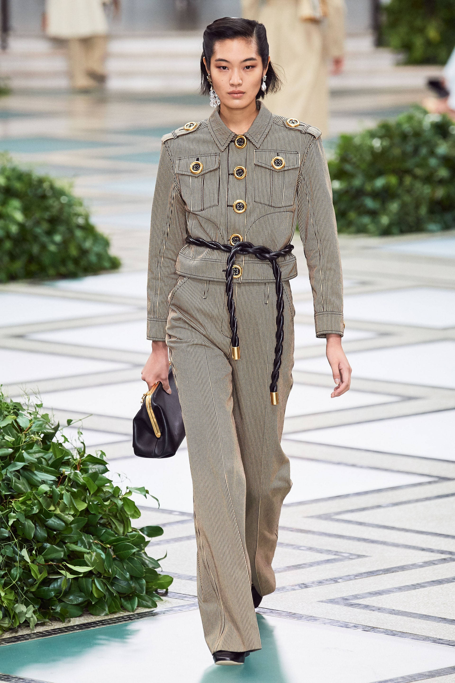 Tory Burch 搭配套裝 壁紙 — 一位模特兒自信地走在天橋上，身穿帶有金色鈕扣和編織腰帶的細條紋軍裝連身褲。