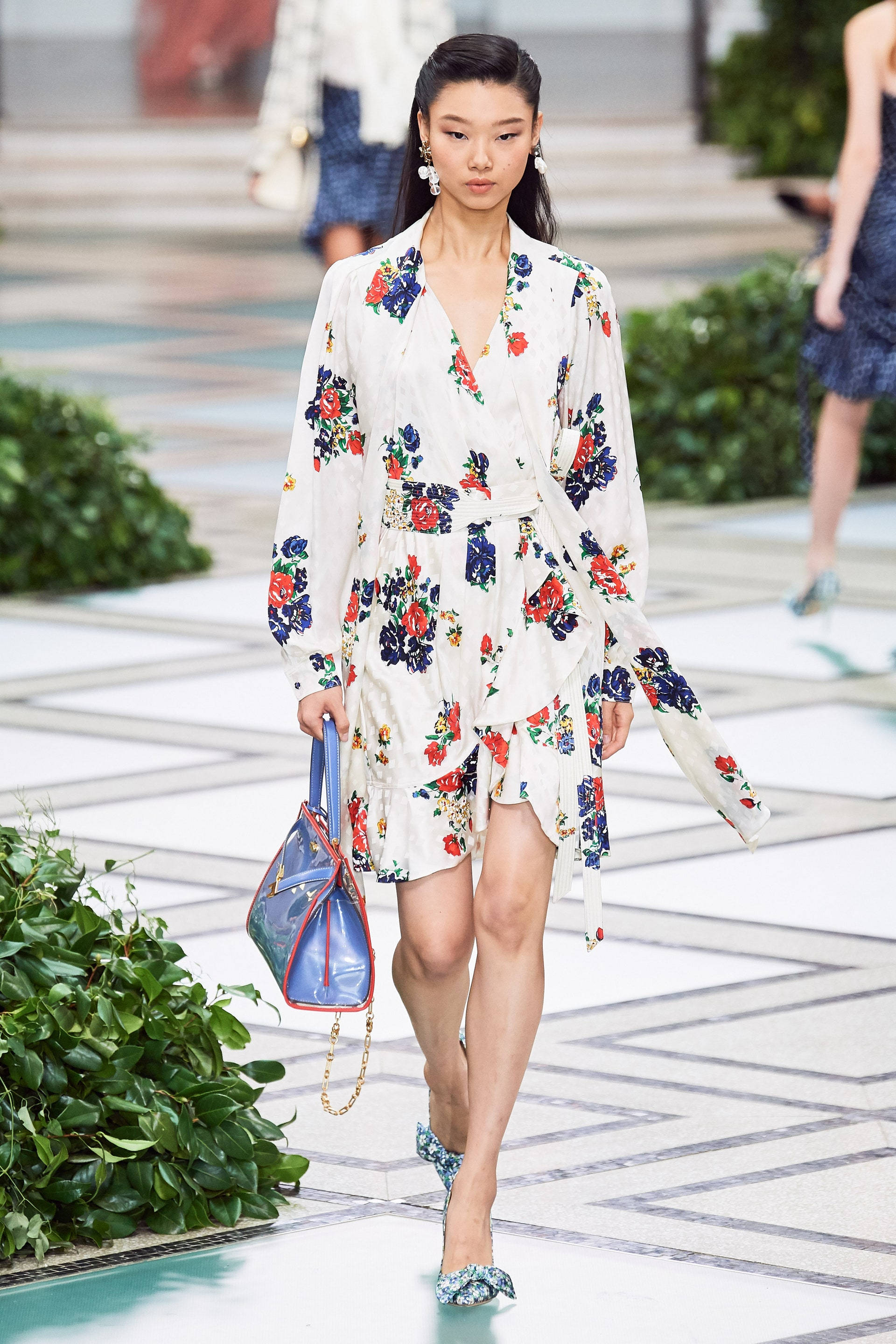Tory Burch 春季時尚 壁紙 — 一位模特兒自信地穿著花卉印花裹身裙和圖案高跟鞋，在幾何瓷磚跑道兩旁綠植的襯托下走秀。