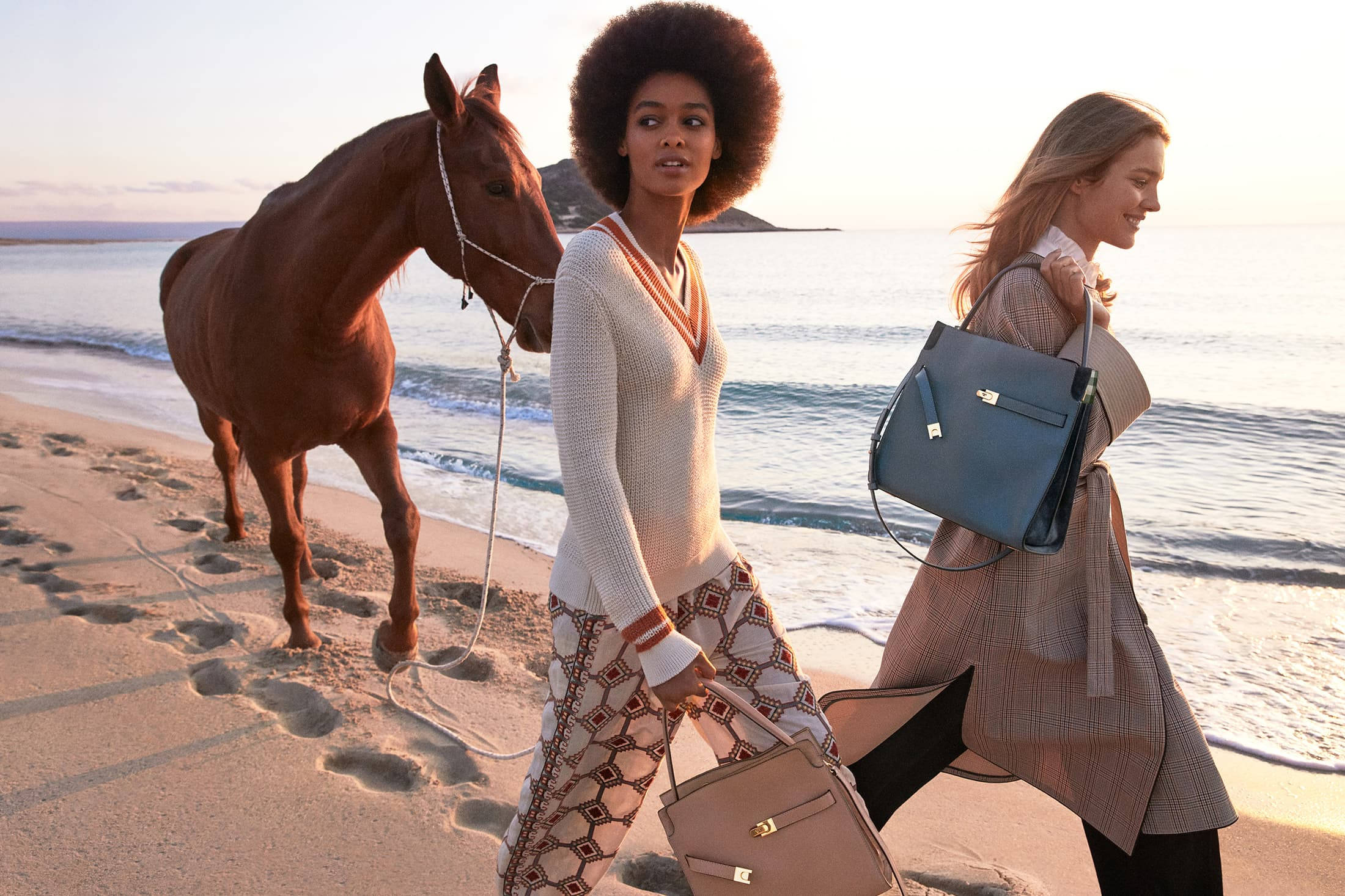 Tory Burch 海濱時尚 壁紙 — 兩位時尚女性在陽光明媚的海灘上漫步，身旁有一匹栗色馬匹，溫柔的海浪輕拍她們的腳。