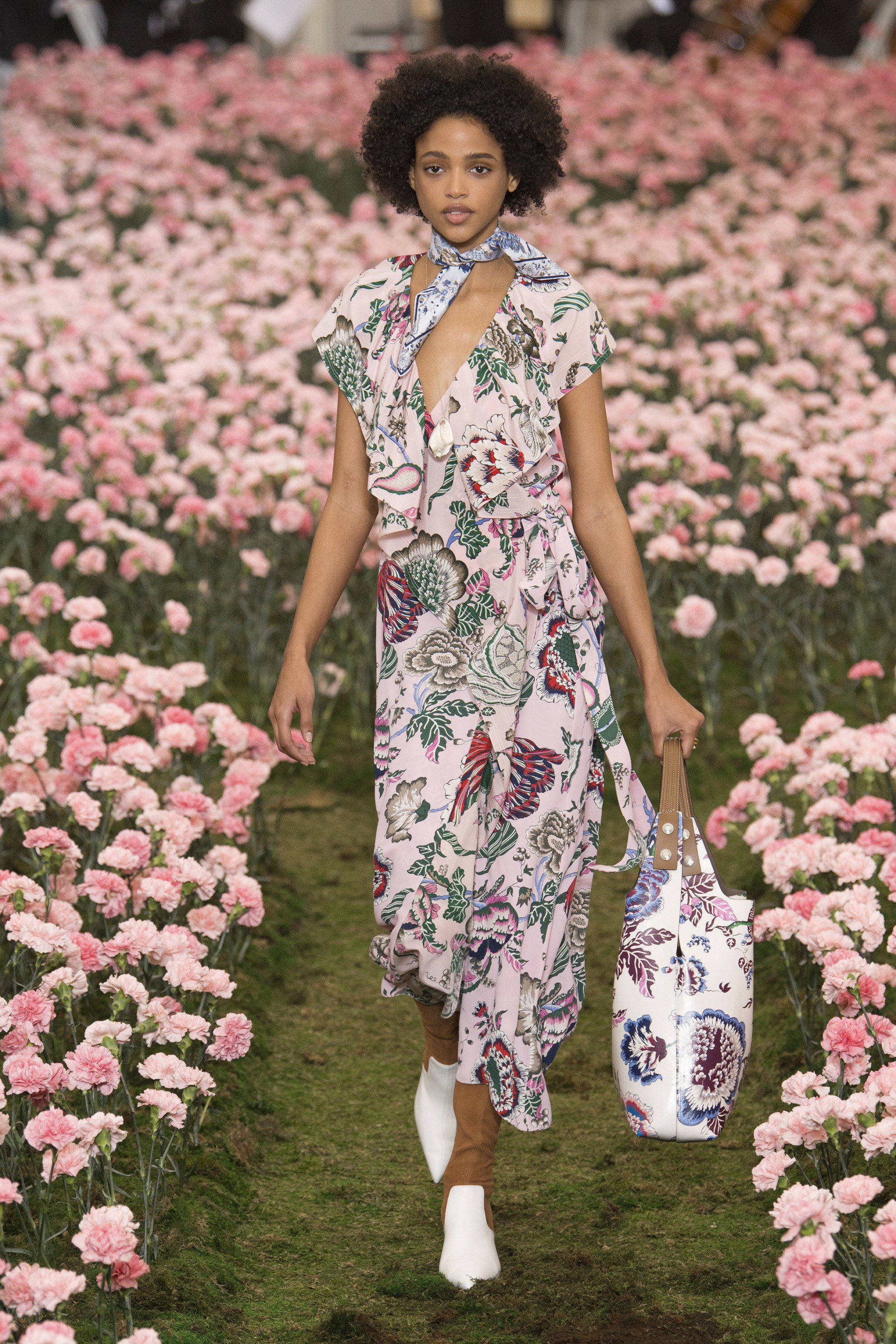 Tory Burch 秋季連衣裙 壁紙 — 一位模特兒自信地穿過粉紅色康乃馨花田，身穿花卉連衣裙並攜帶相配的手袋。