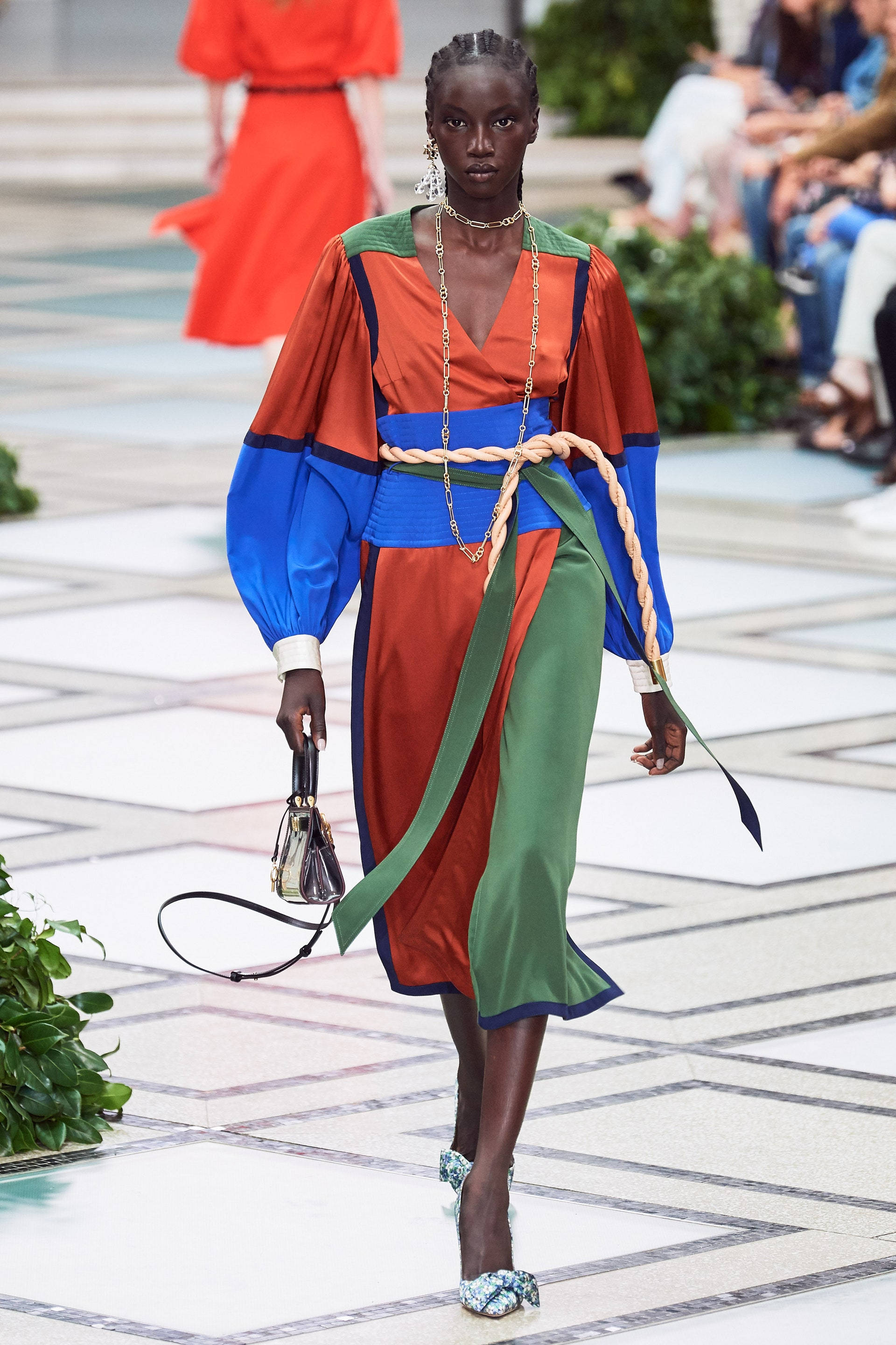 Tory Burch 色彩繽紛連衣裙 壁紙 — 一位模特兒自信地走在瓷磚跑道上，身穿大膽的橙色、藍色和綠色拼色連衣裙，搭配編織繩腰帶和圖案高跟鞋。