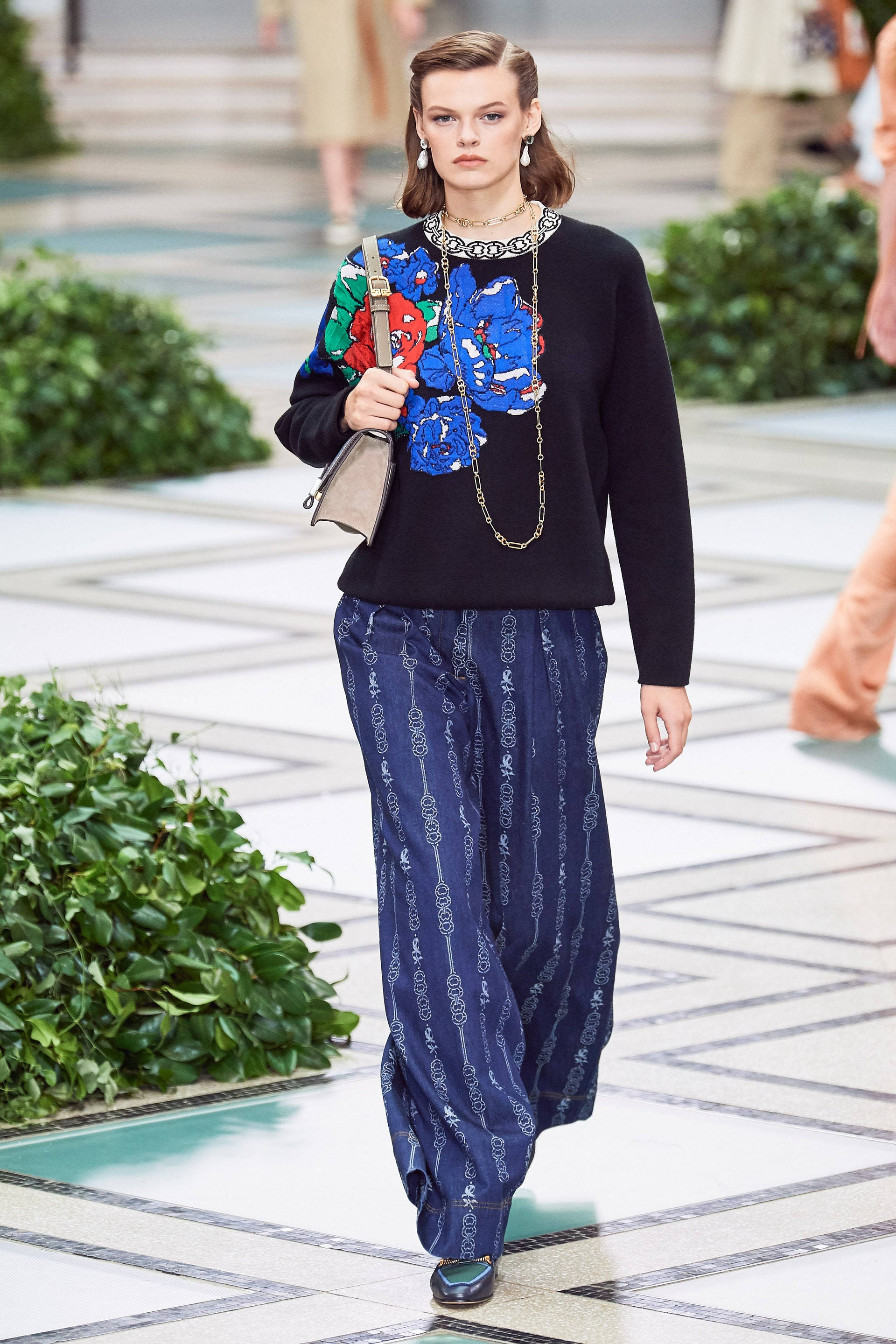 Tory Burch 跑秀時尚 壁紙 — 一位模特兒自信地走在瓷磚跑道上，兩旁是綠色樹籬，身穿黑色毛衣，帶有鮮明的藍色花卉刺繡和圖案藍色褲子。