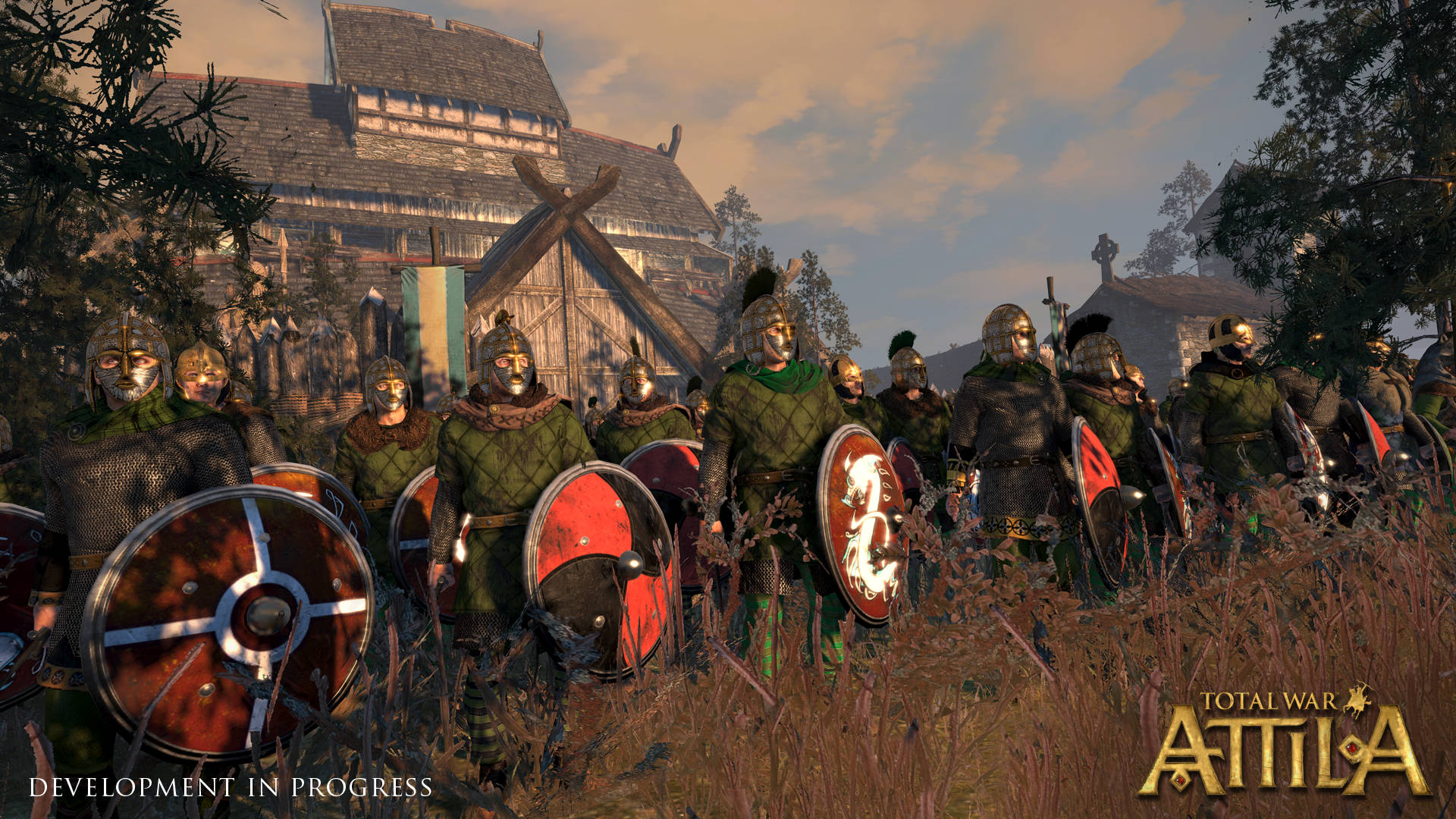 Total War Attila 格鬥士整齊列隊 壁紙 — 一支穿著鎖子甲和頭盔的中世紀軍隊，在日落時分，於木建築附近的草地上，手持圓盾列陣。