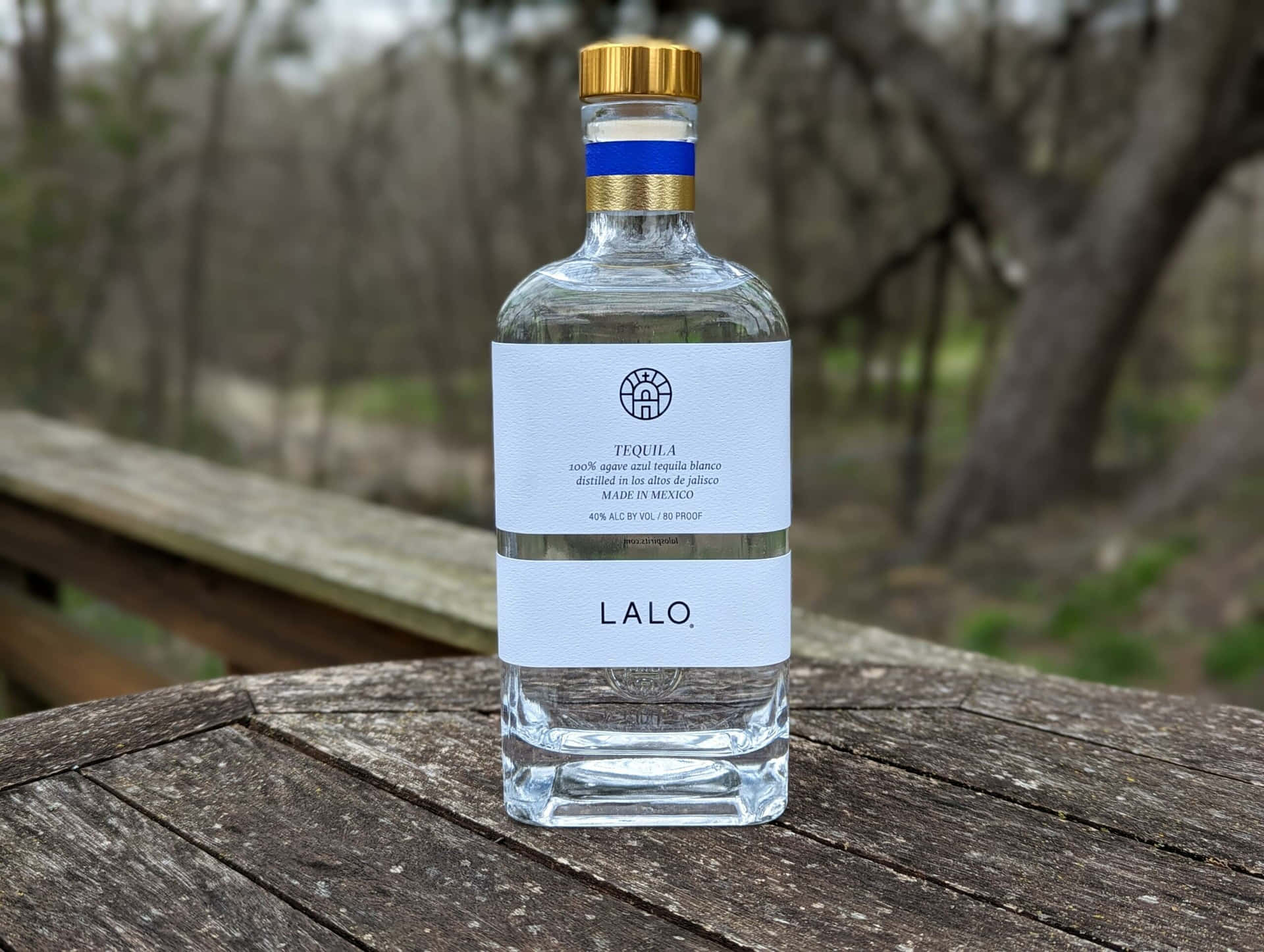  壁紙 — 一個透明嘅Lalo龍舌蘭酒樽，金色樽蓋，擺喺室外一張風化咗嘅木枱上面，背景有模糊嘅樹木。