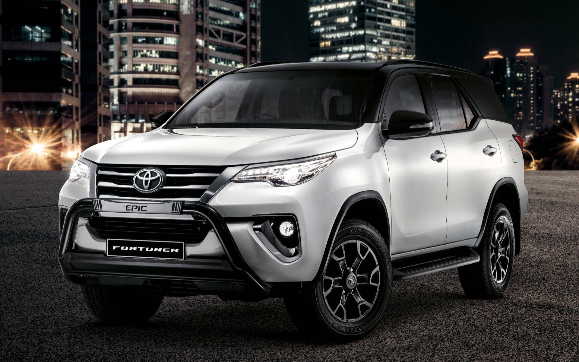 至尊白色 Toyota Fortuner — 奢華與科技的完美結合 壁紙 — 一輛帶有黑色飾邊的銀色豐田 Fortuner SUV 在夜間城市景觀中醒目地停放，背後是燈火通明的摩天大樓。