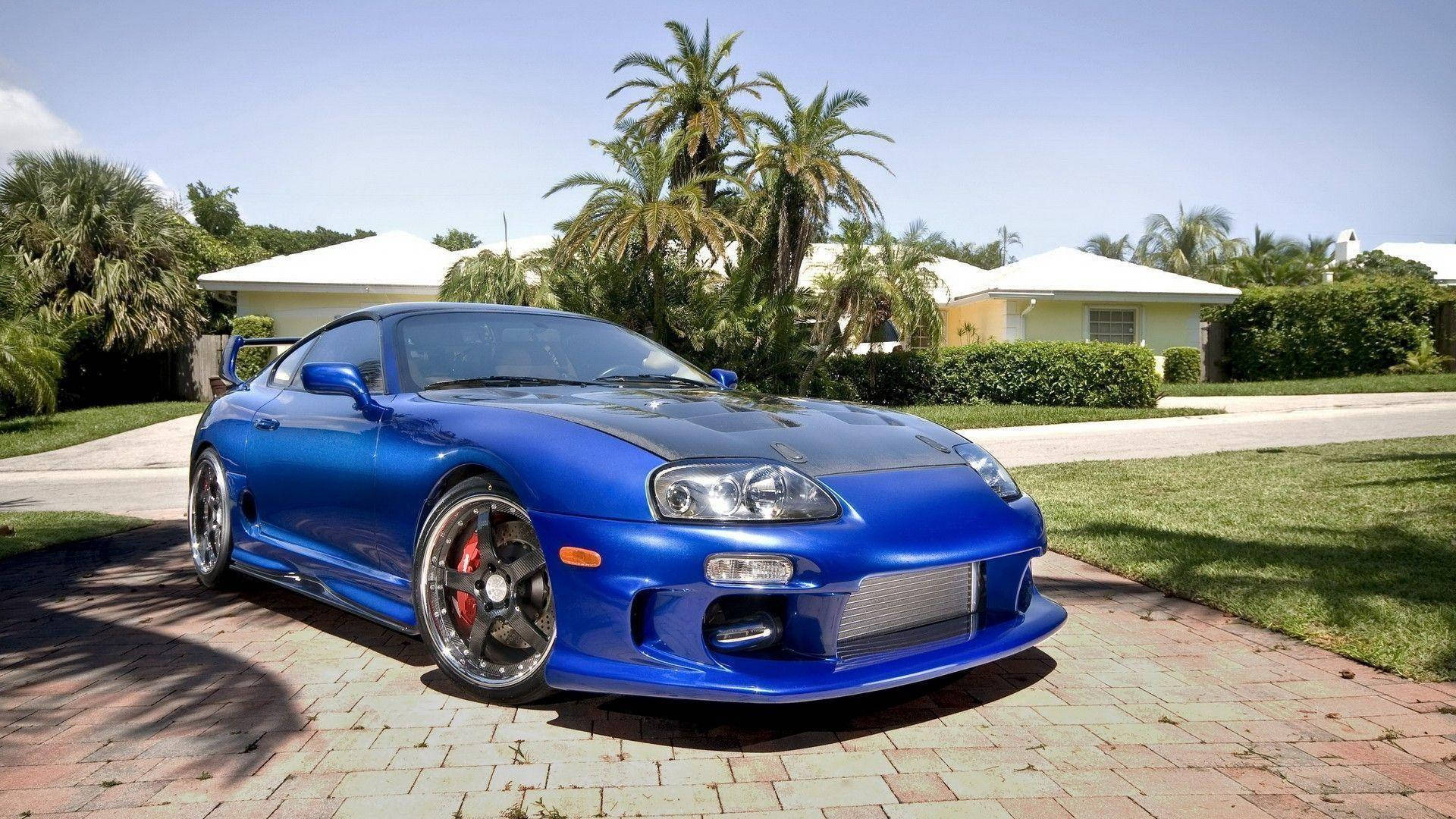 藍色Toyota Supra Mk4碳纖維引擎蓋 壁紙 — 一輛充滿活力的藍色豐田Supra跑車停在磚砌車道上，背景有棕櫚樹和郊區房屋，天空晴朗。