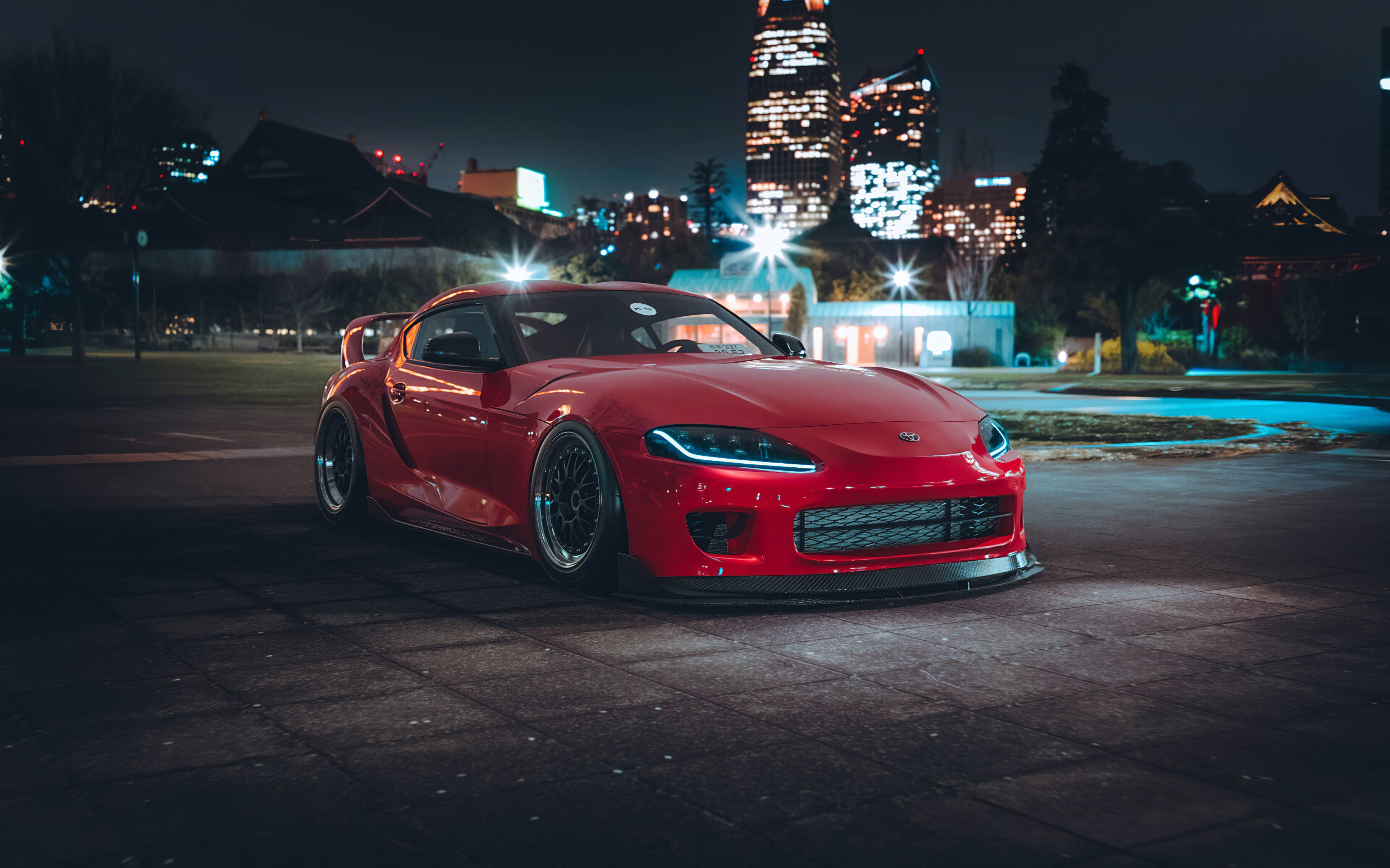 紅色Toyota Supra停泊城市夜景 壁紙 — 一輛鮮紅色的跑車在夜晚的城市燈光下，映襯著黑暗的城市天際線閃閃發光。