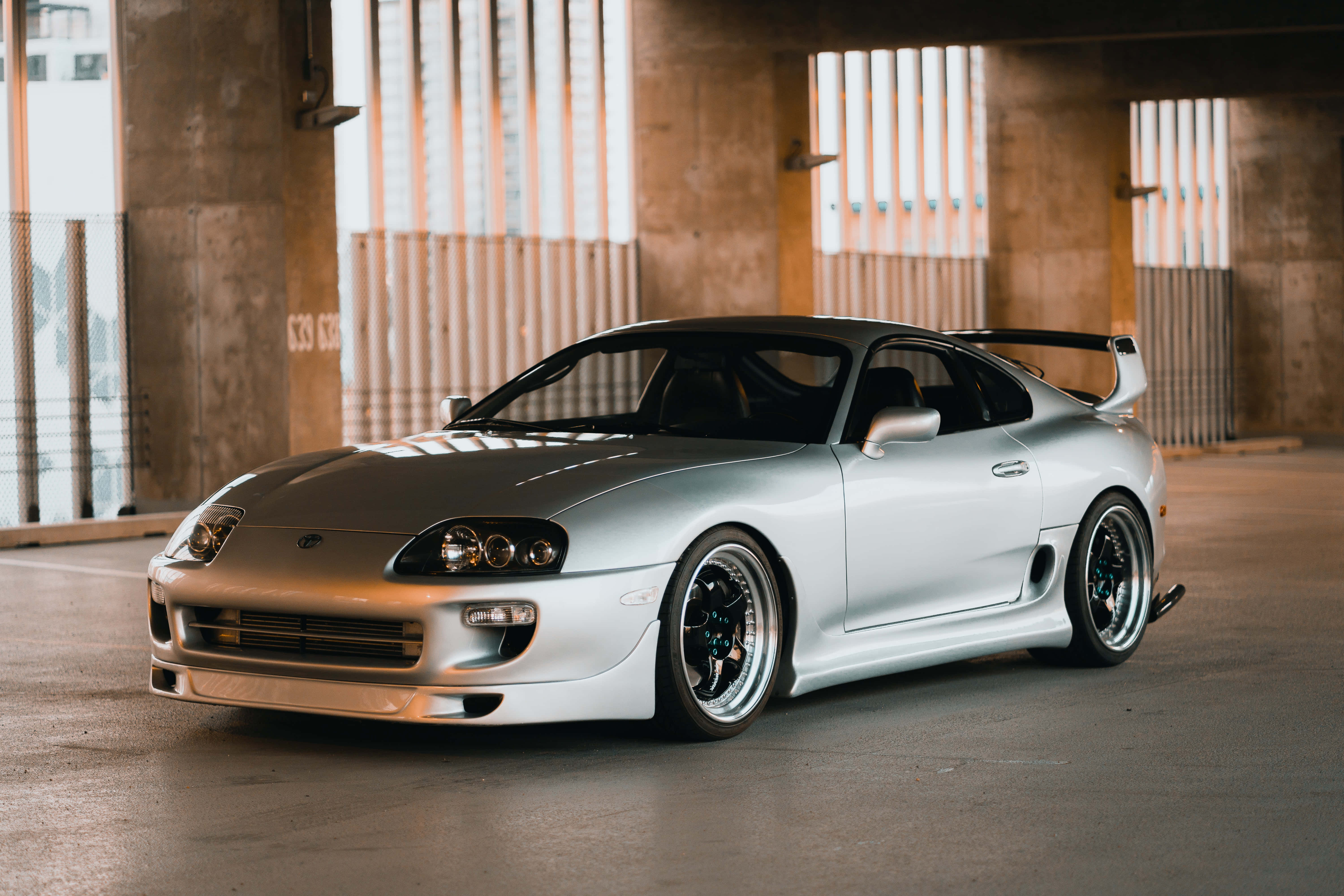 以時尚姿態駕馭 Toyota Supra 壁紙 — 一架時尚的銀色豐田Supra跑車，帶有後擾流板，停放在一個帶有垂直百葉窗的混凝土天橋下，讓柔和的光線透入。
