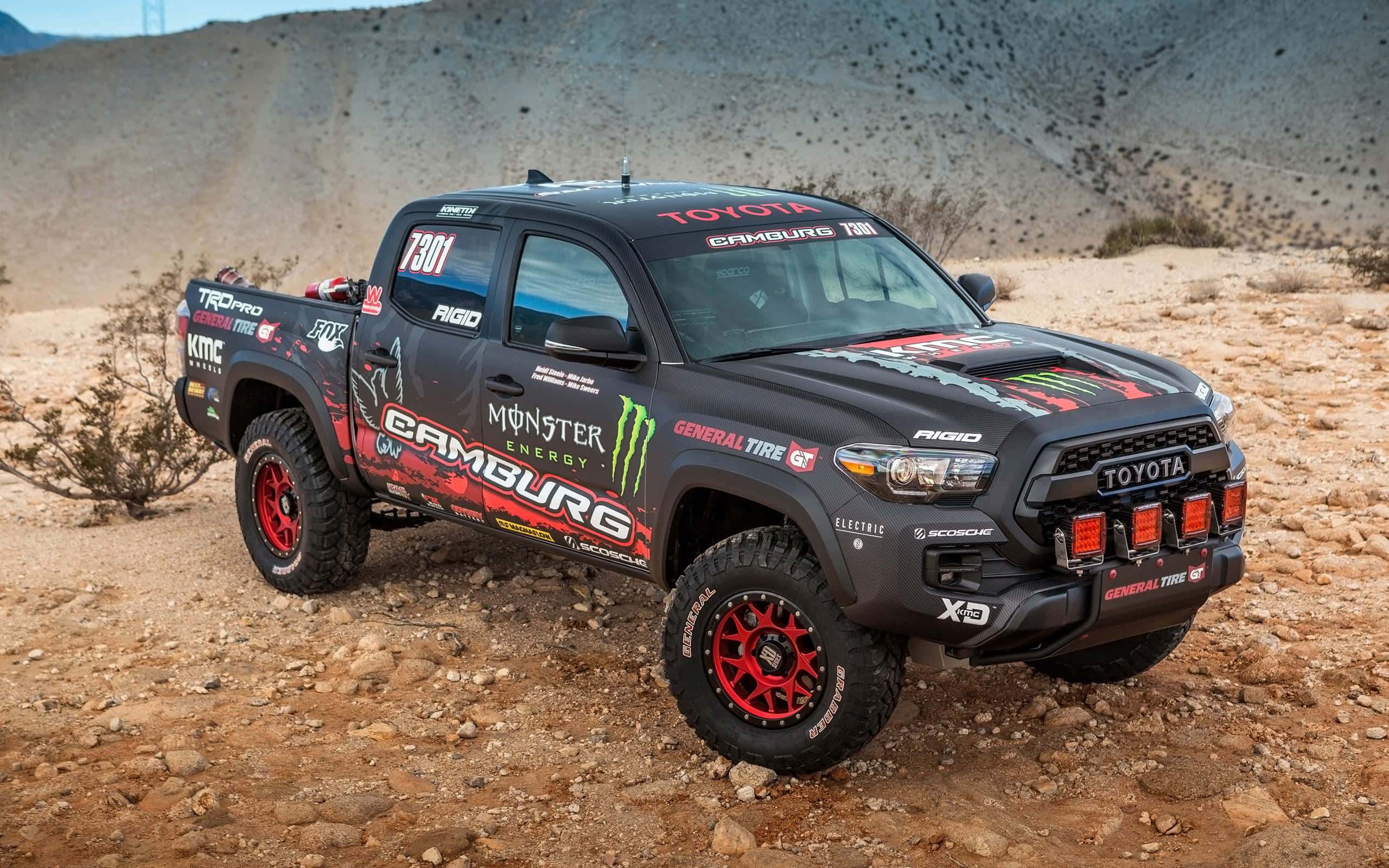 Toyota TRD 定制 Tacoma 壁紙 — 一輛帶有紅色點綴和Monster Energy貼紙的黑色豐田Tacoma賽車卡車在岩石沙漠地形中行駛。