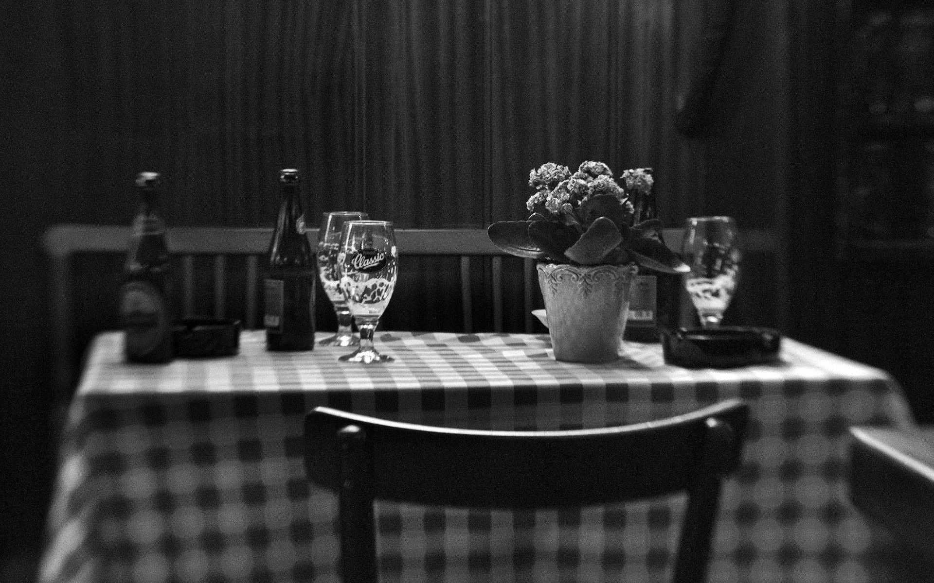 Trattoria Da Fausto 美式高雅用餐體驗 壁紙 — 一張黑白照片，展示一張質樸的桌子，上面放著啤酒瓶、玻璃杯和一盆植物，光線昏暗。