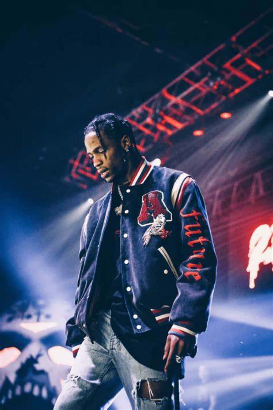 在趣味的Travis Scott iPhone上享受音樂與影片 壁紙 — 一位身穿海軍藍校隊夾克的饒舌歌手站在燈光昏暗的舞台上，沐浴在紅色和藍色的聚光燈下，煙霧在他周圍旋轉。