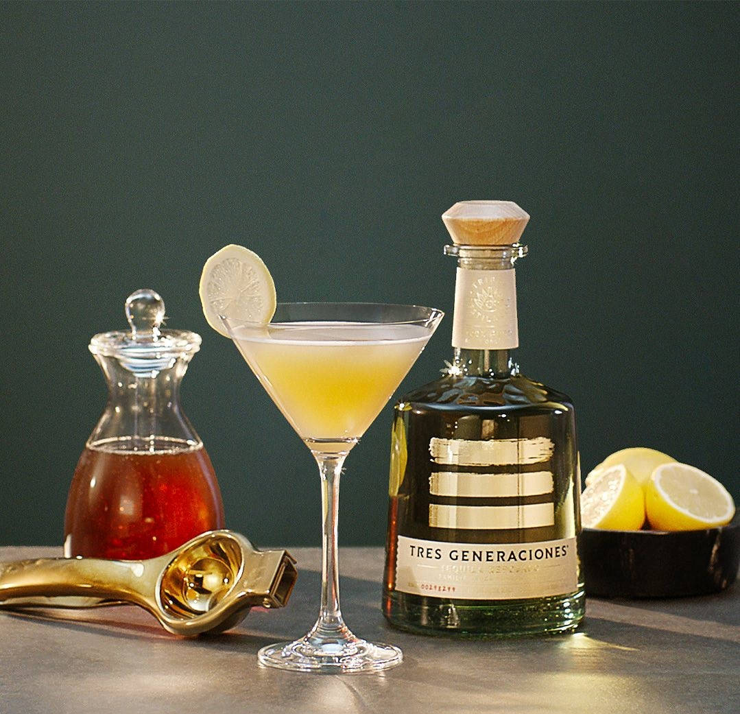 Tres Generaciones 鸡尾酒飲品 壁紙 — 一杯金色雞尾酒盛載於馬提尼杯，飾以檸檬片，旁邊放著一瓶Tres Generaciones龍舌蘭酒和一碗檸檬，置於深色桌上。