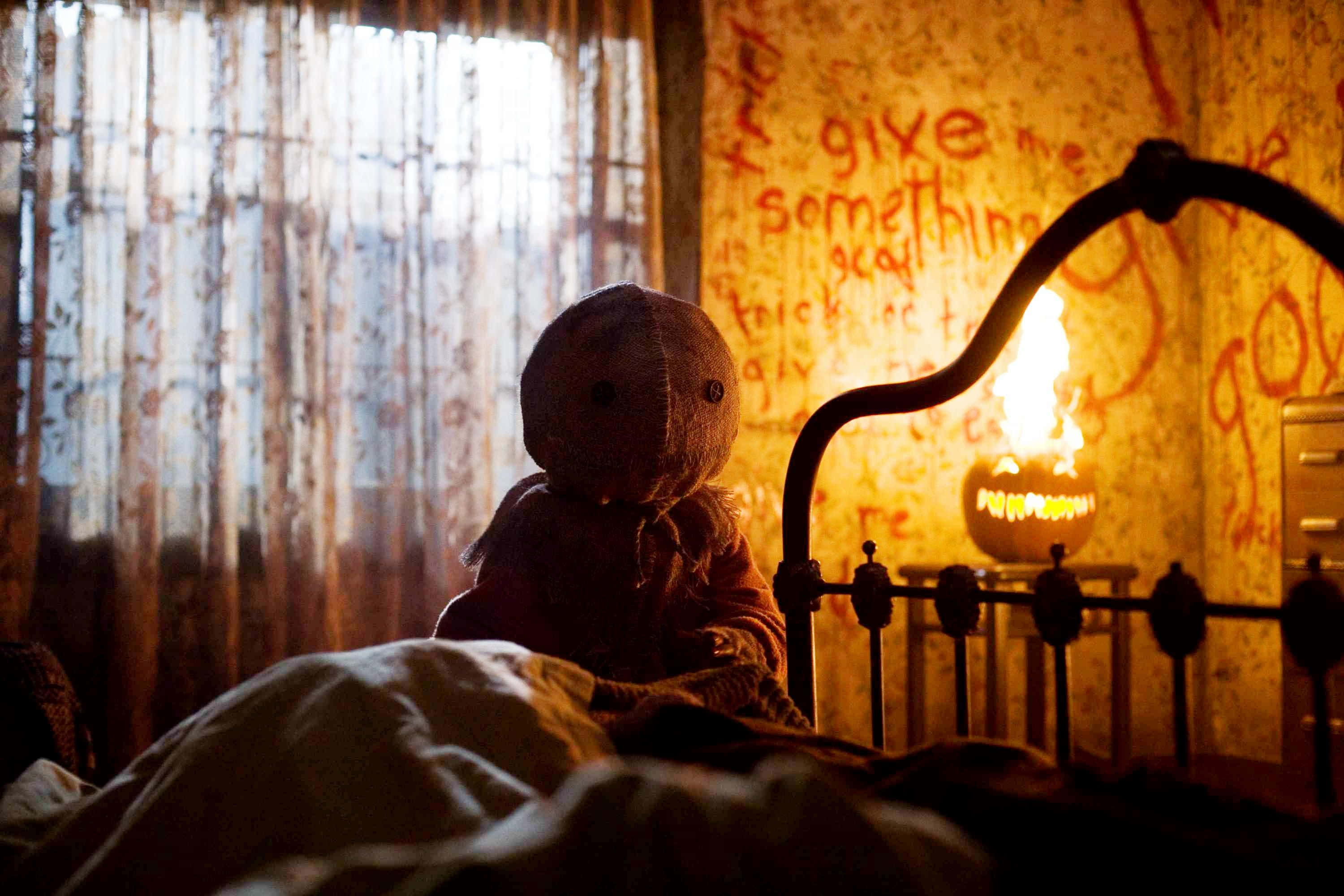 Trick 'r Treat 中的 Sam 躺在床上 壁紙 — 一個恐怖的手工娃娃坐在燈光昏暗的床上，旁邊有閃爍的南瓜燈和牆上不祥的紅色塗鴉。