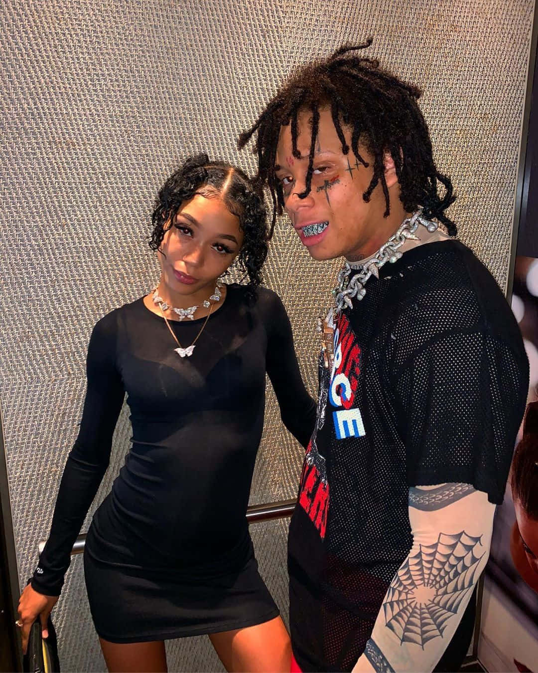 Trippie Redd 與 Coi Leray 的戀愛模範照片 壁紙 — 兩位穿著時尚的人士自信地擺姿勢，背景是帶有紋理的金屬牆，配以大膽的珠寶和紋身。