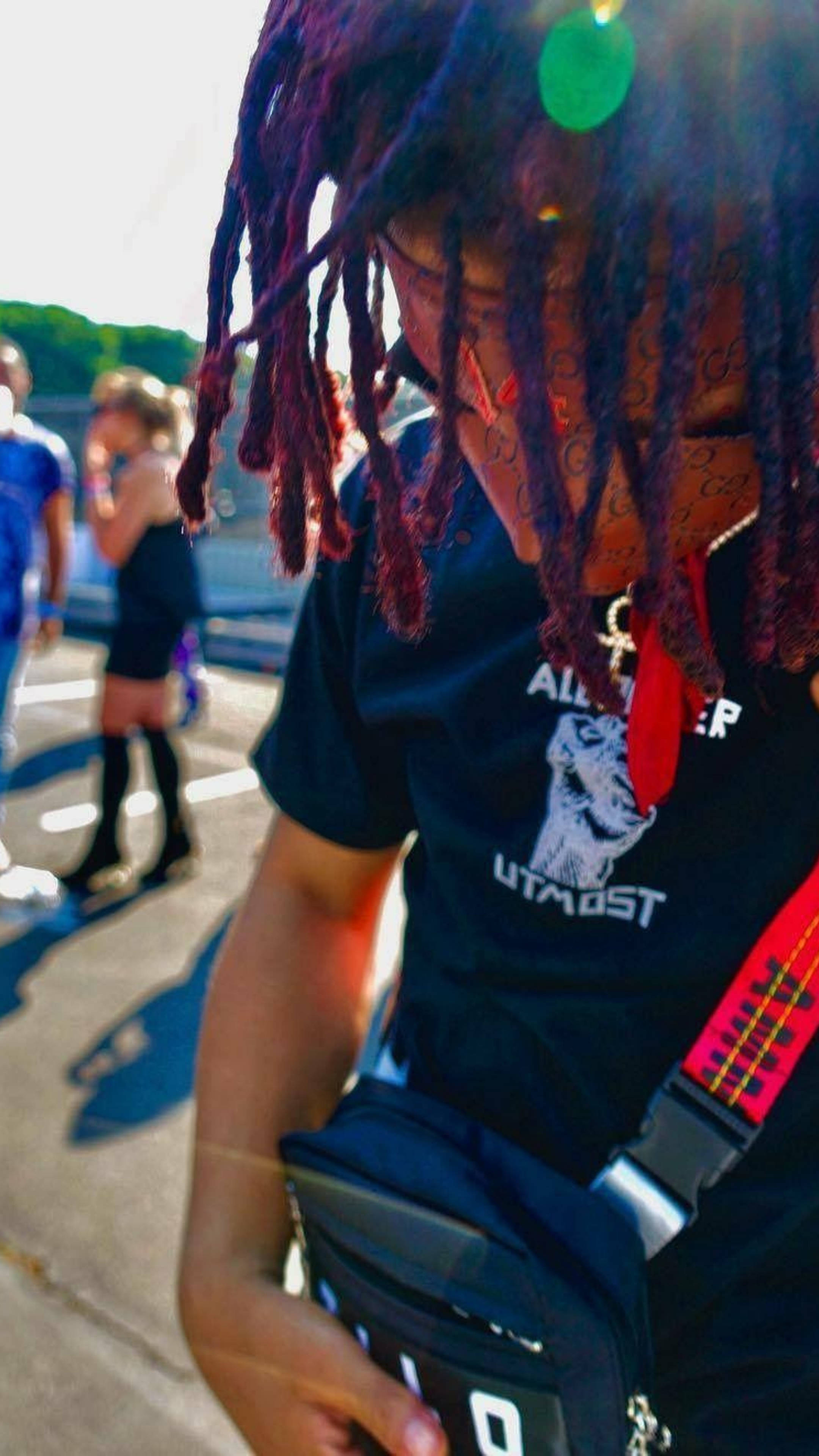 Trippie Redd 偷拍瞬間 壁紙 — 特寫：一個人戴著鮮紫色辮子頭和口罩，站在戶外明亮的陽光下。