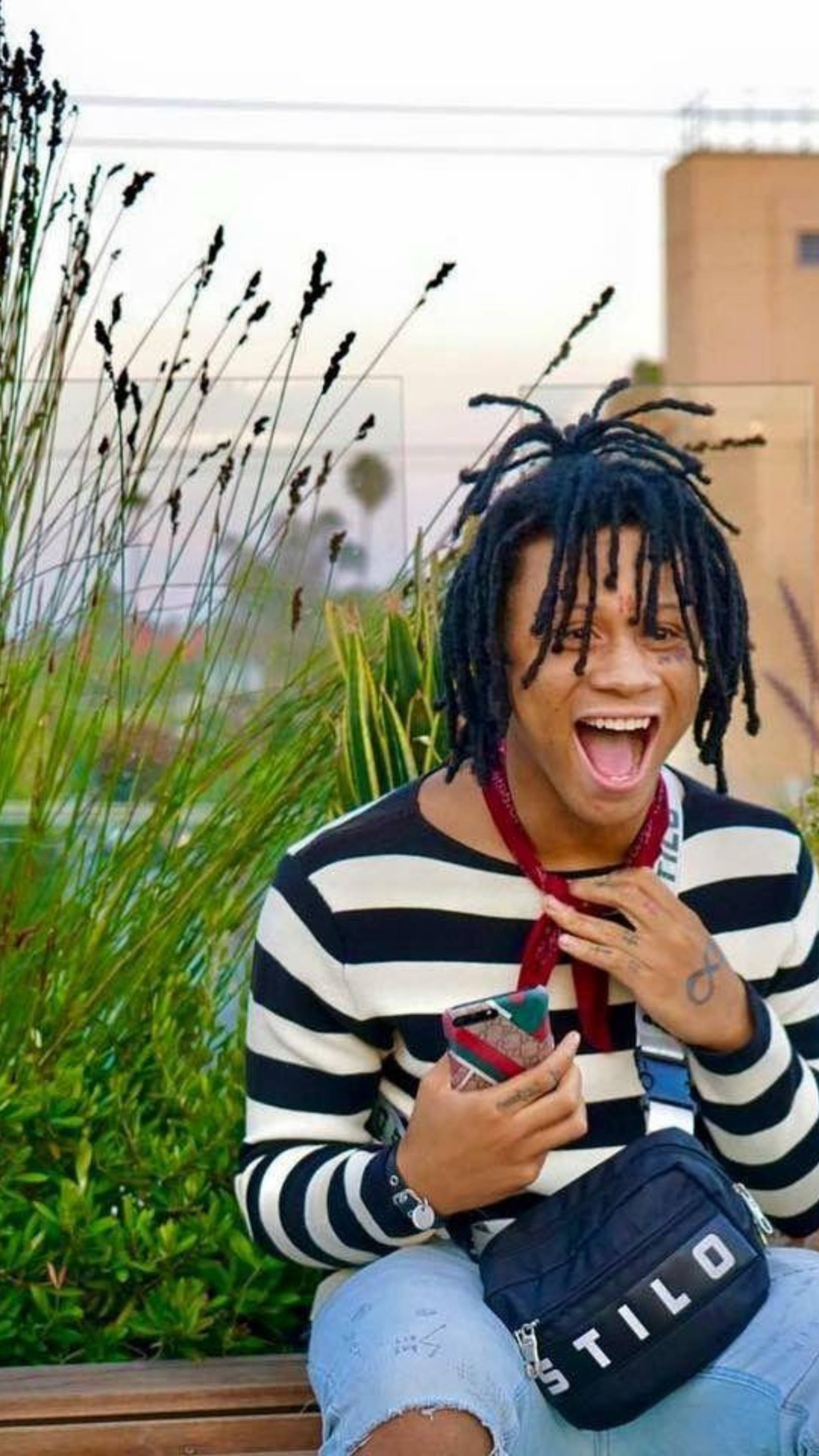 Trippie Redd 條紋造型 壁紙 — 一個快樂的年輕黑人辮子男坐在長椅上，拿著電話和腰包大笑。