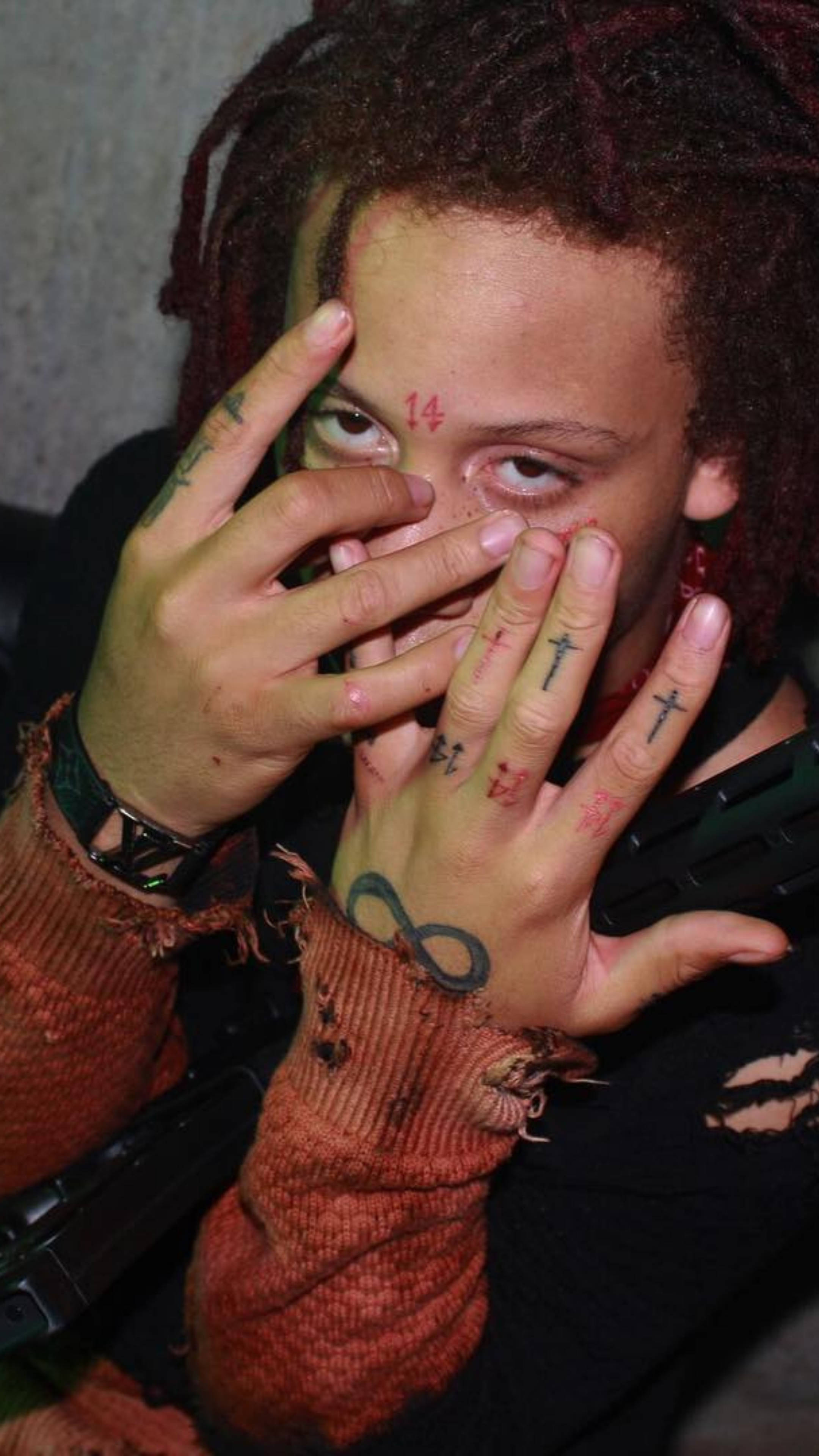Trippie Redd 無限紋身 壁紙 — 一個有紅色辮子頭和手部及面部紋身的人，在昏暗的環境中用手指部分遮蓋面部。