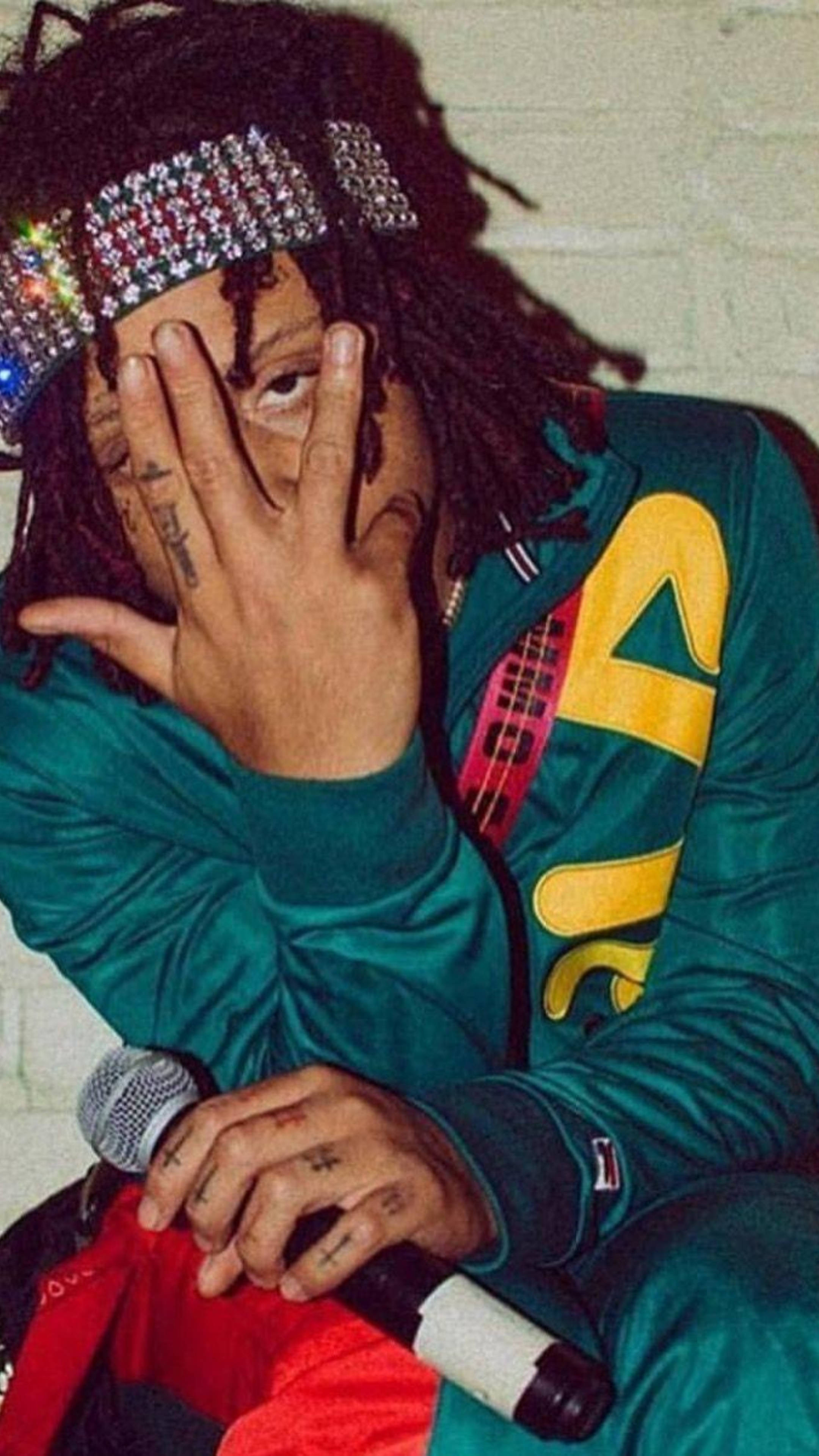 Trippie Redd 獨特肖像 壁紙 — 一個有辮子頭和鑲寶石頭帶的人，部分遮住臉，同時拿著麥克風靠著磚牆。