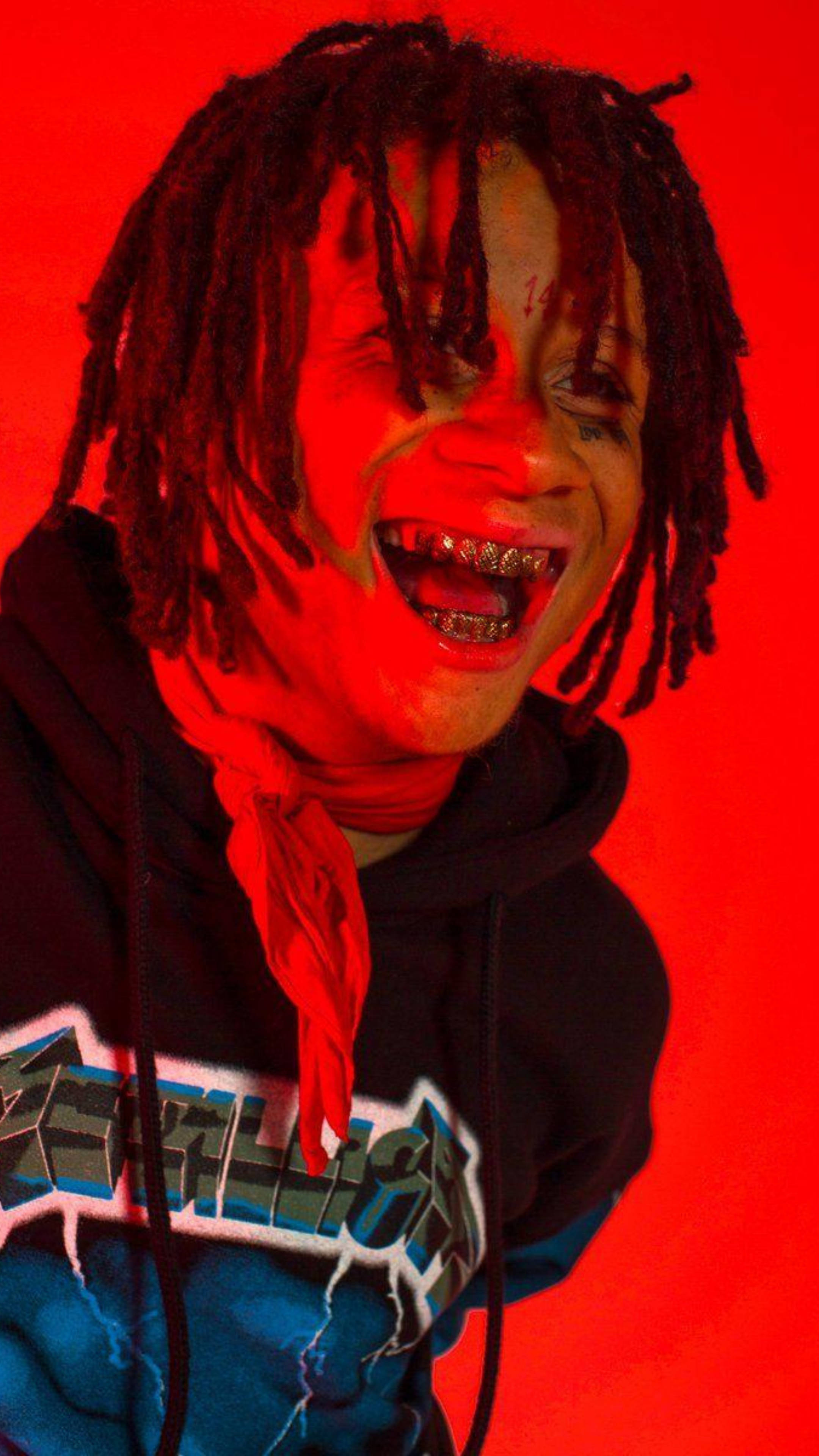 Trippie Redd 笑容滿面 壁紙 — 一個留著辮子頭、戴著金牙套的人，在鮮紅色背景下穿著帶有圖案設計的黑色連帽衫，大笑不止。