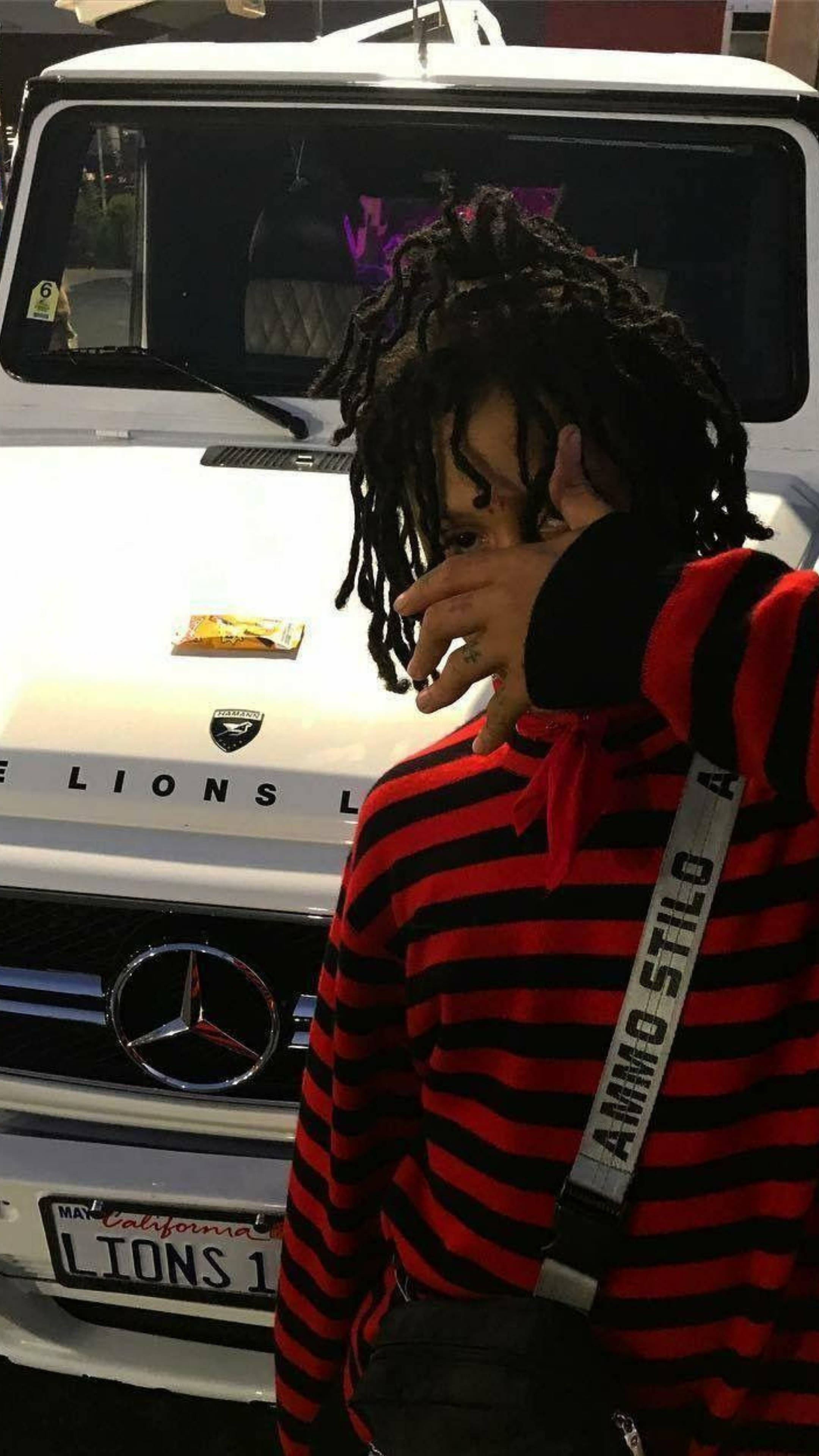 Trippie Redd 編織毛衣 壁紙 — 一個留著辮子的人穿著紅黑條紋連帽衫，部分遮住臉，站在一輛白色Mercedes-Benz G-Wagen前。