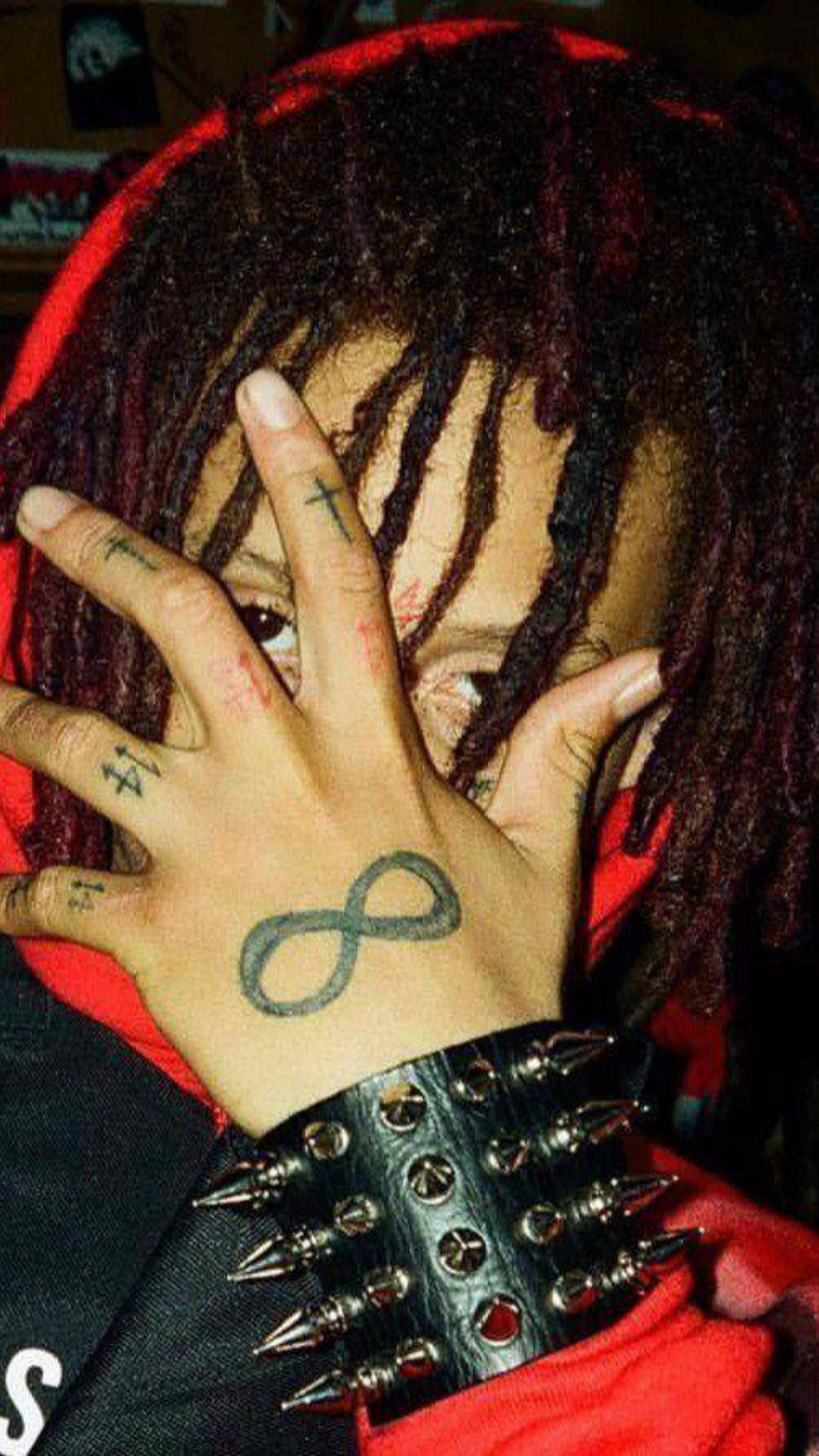 Trippie Redd 鬼馬腕帶 壁紙 — 一個有紅色辮子和帶刺腕帶的人，用手部分遮住臉，手上顯示包括無限符號的紋身。