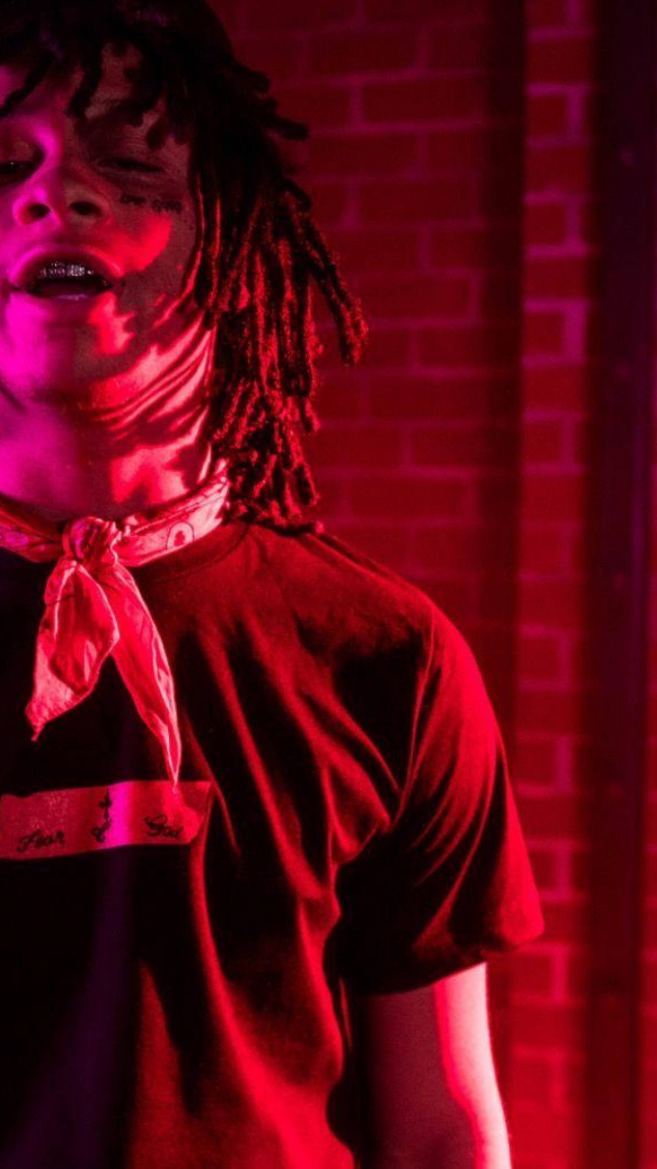 Trippie Redd 黑色印花上衣 壁紙 — 一個留著辮子、戴著紅色頭巾的人站在磚牆前，沐浴在強烈的紅色和洋紅色燈光中。