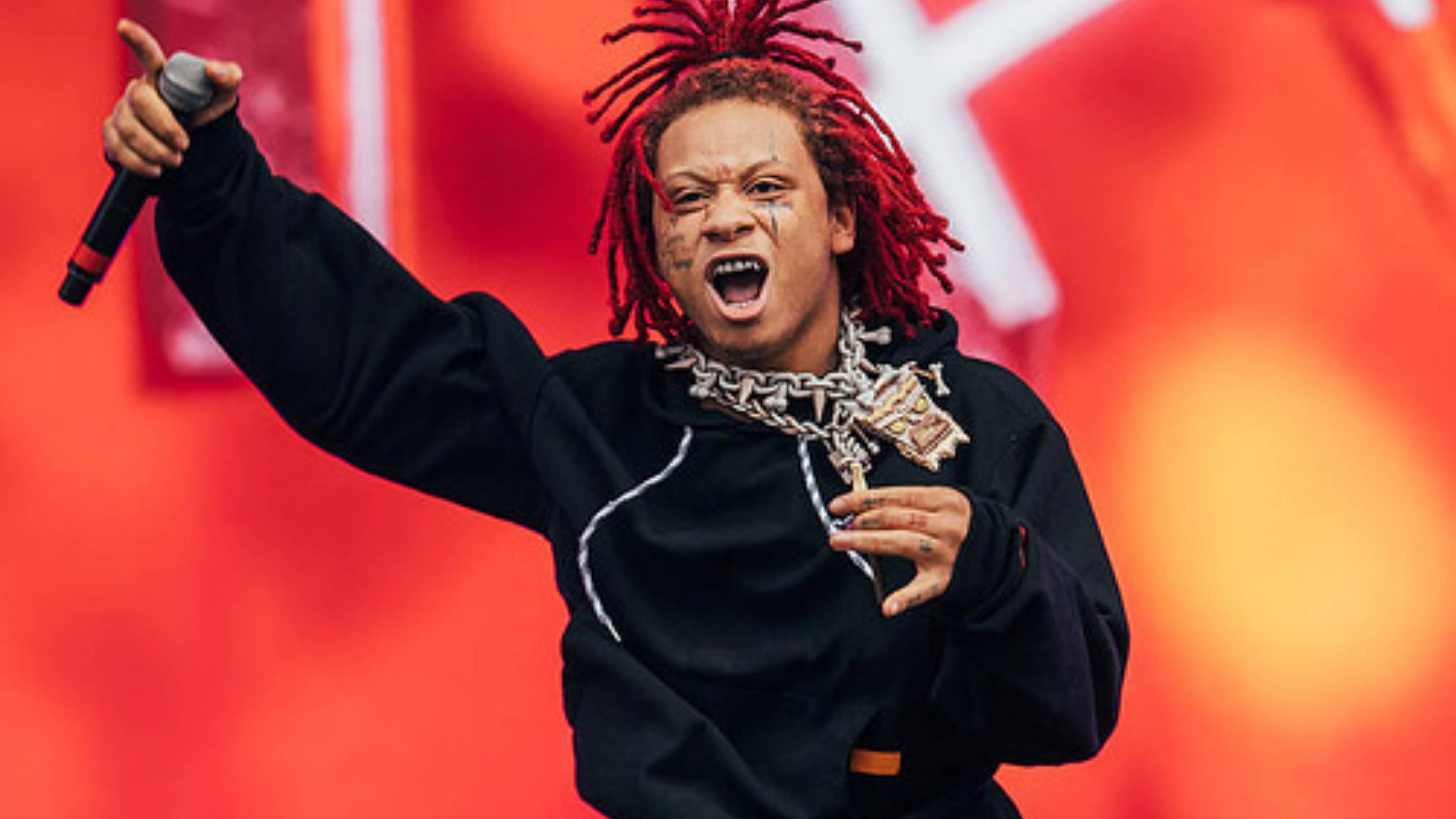 Trippie Redd 黑色連帽衫 壁紙 — 一位擁有紅色辮子頭和金鏈的饒舌歌手在舞台上熱情演出，手持麥克風，背景是鮮紅色的。