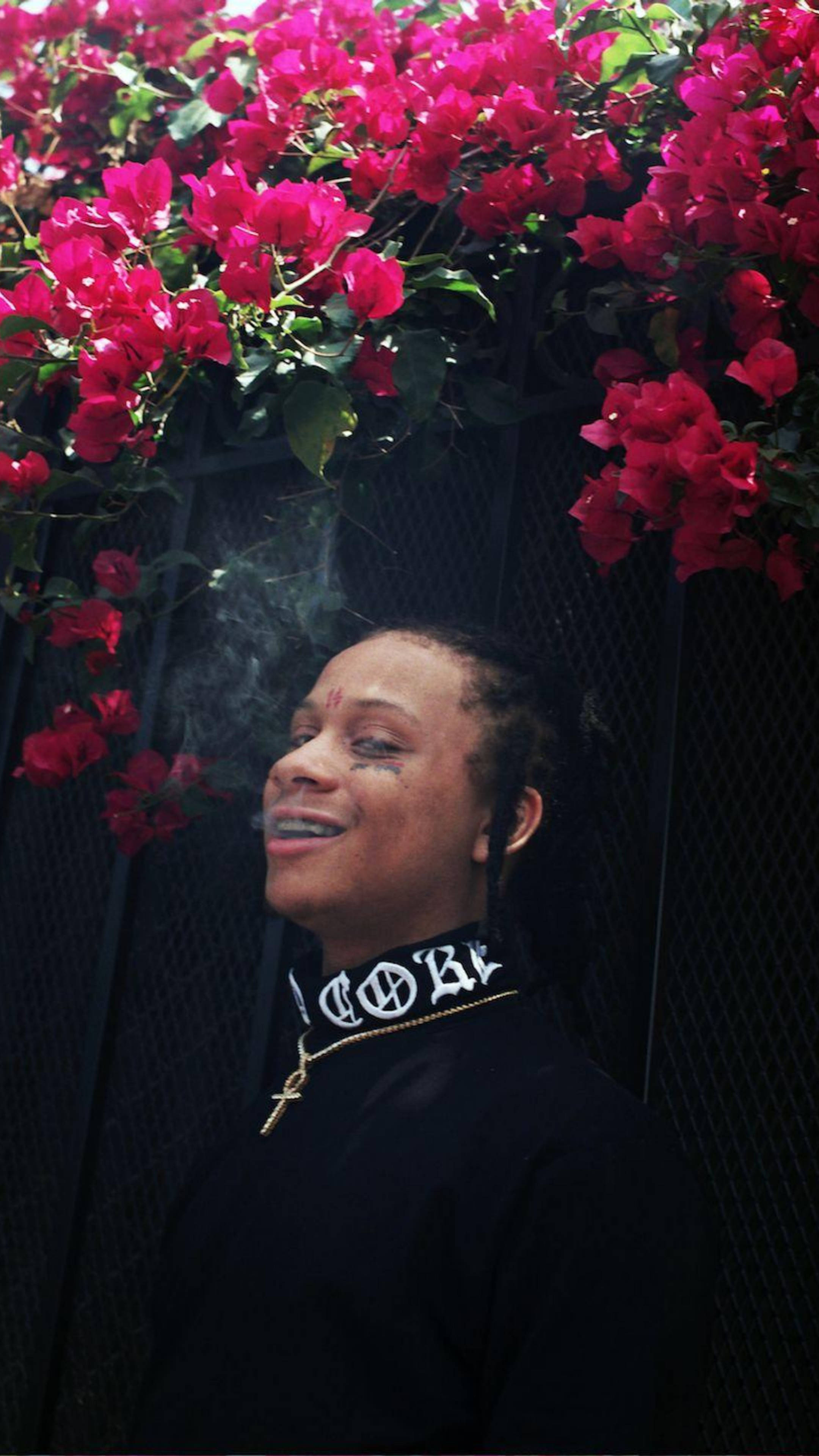 Trippie Redd 黑色高領毛衣 壁紙 — 一個帶著喜悅表情的人，在深色網格圍欄前，於一串串鮮豔粉紅色的三角梅瀑布下吸煙。