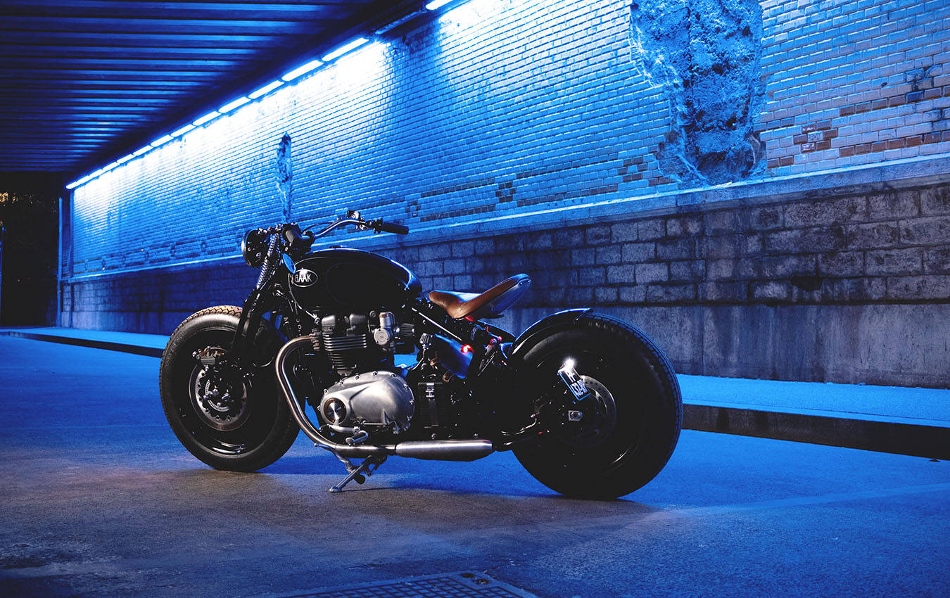 Triumph Bonneville Moon Bobber 1200 月光下 壁紙 — 一架時尚黑色訂製電單車配備厚輪胎，停放在藍色燈光隧道內，牆壁為磚牆。