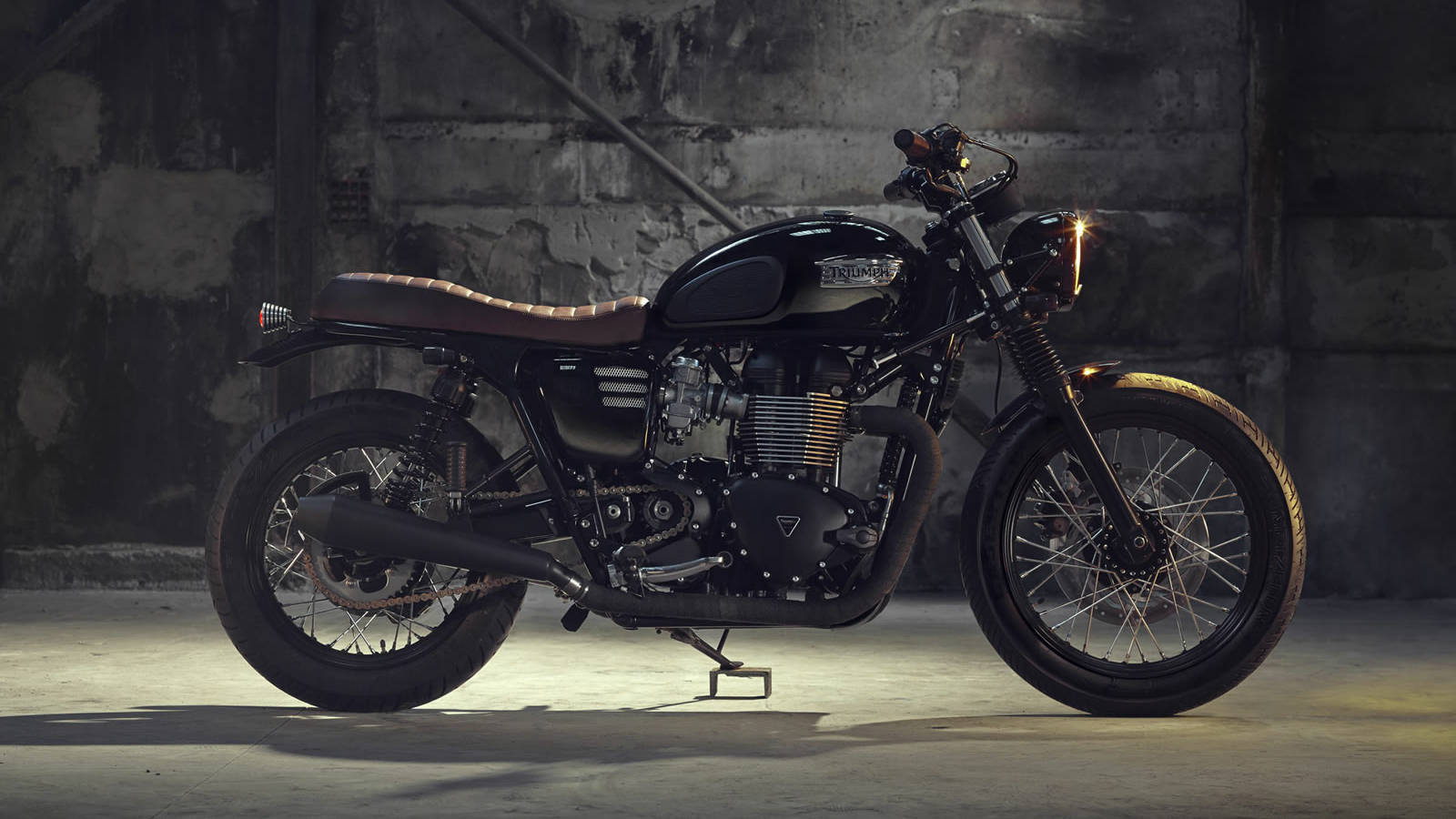 Triumph Bonneville T100 機車 壁紙 — 一架型格黑色Triumph電單車配棕色皮革座椅，在燈光昏暗的混凝土車庫中，被聚光燈戲劇性地照亮。
