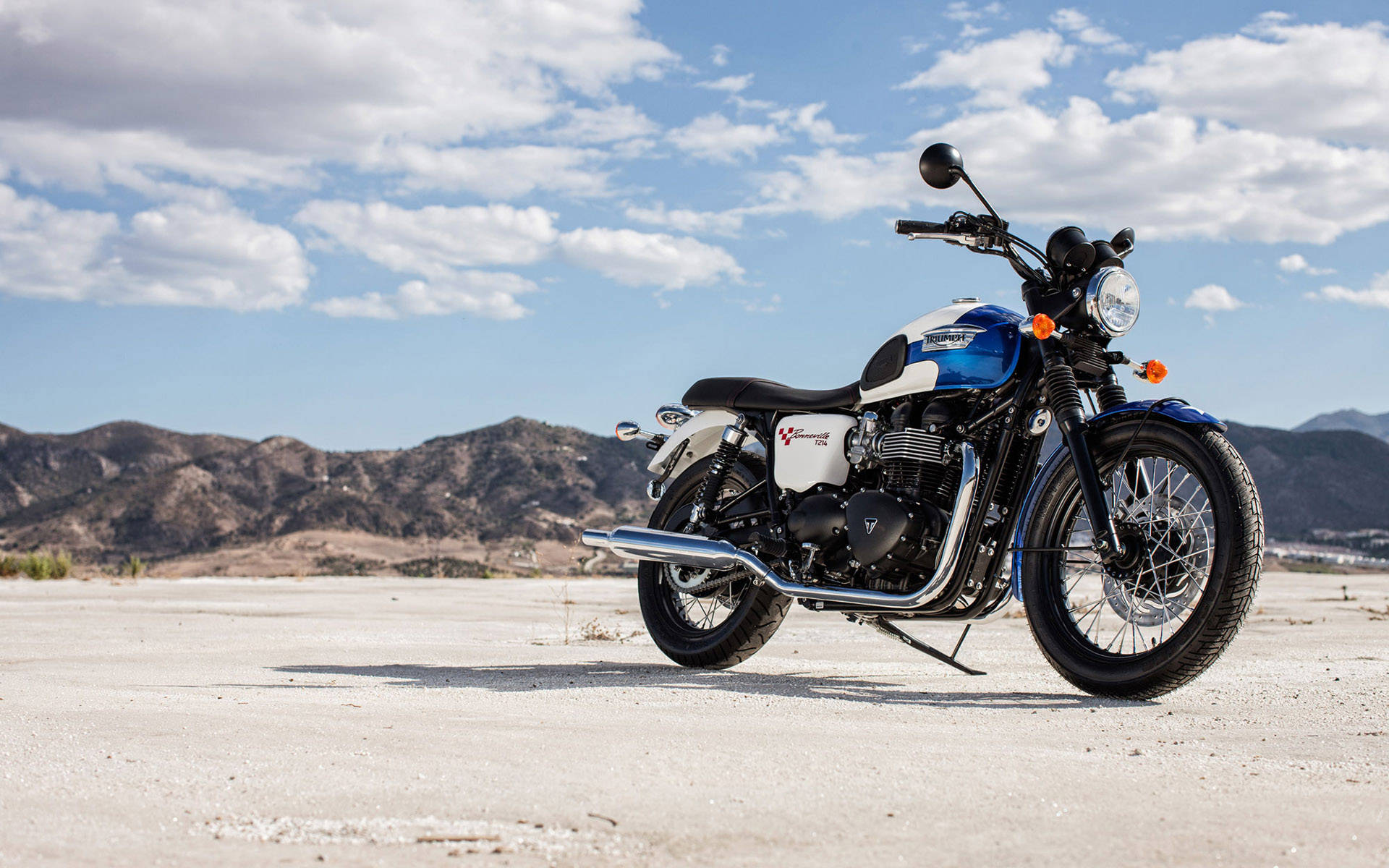 Triumph Bonneville 2015 藍色 Newchurch SE 壁紙 — 一架藍白色經典電單車獨自停喺陽光照耀嘅沙漠平原上，遠處有山脈，天空局部多雲。