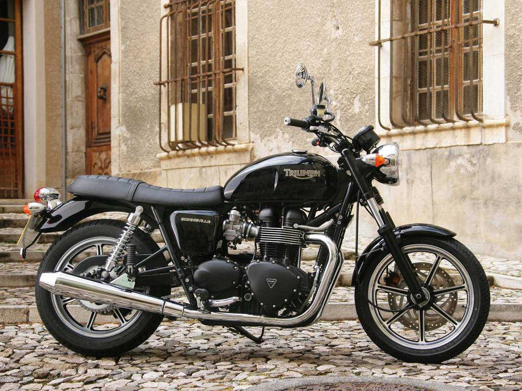 Triumph Bonneville T120 摩托車 壁紙 — 一架時尚的黑色 Triumph Bonneville 電單車停泊在陽光普照的鵝卵石街道上，旁邊是一座風化的石建築，帶有鐵欄窗戶。