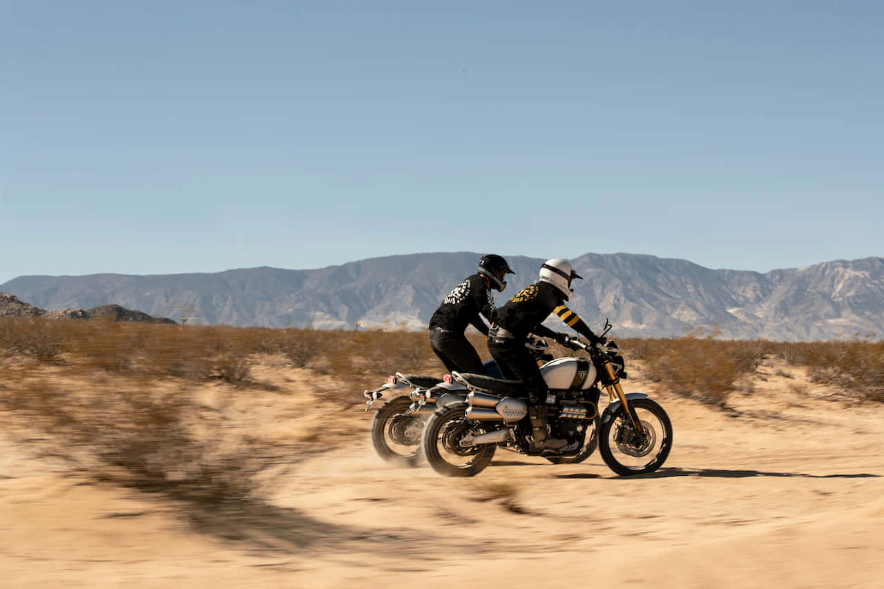 Triumph Scrambler最佳自行車沙漠冒險背景 壁紙 — 兩名電單車手在烈日曝曬的沙漠上競速，揚起塵土，遠處是崎嶇的山脈，天空清澈湛藍。