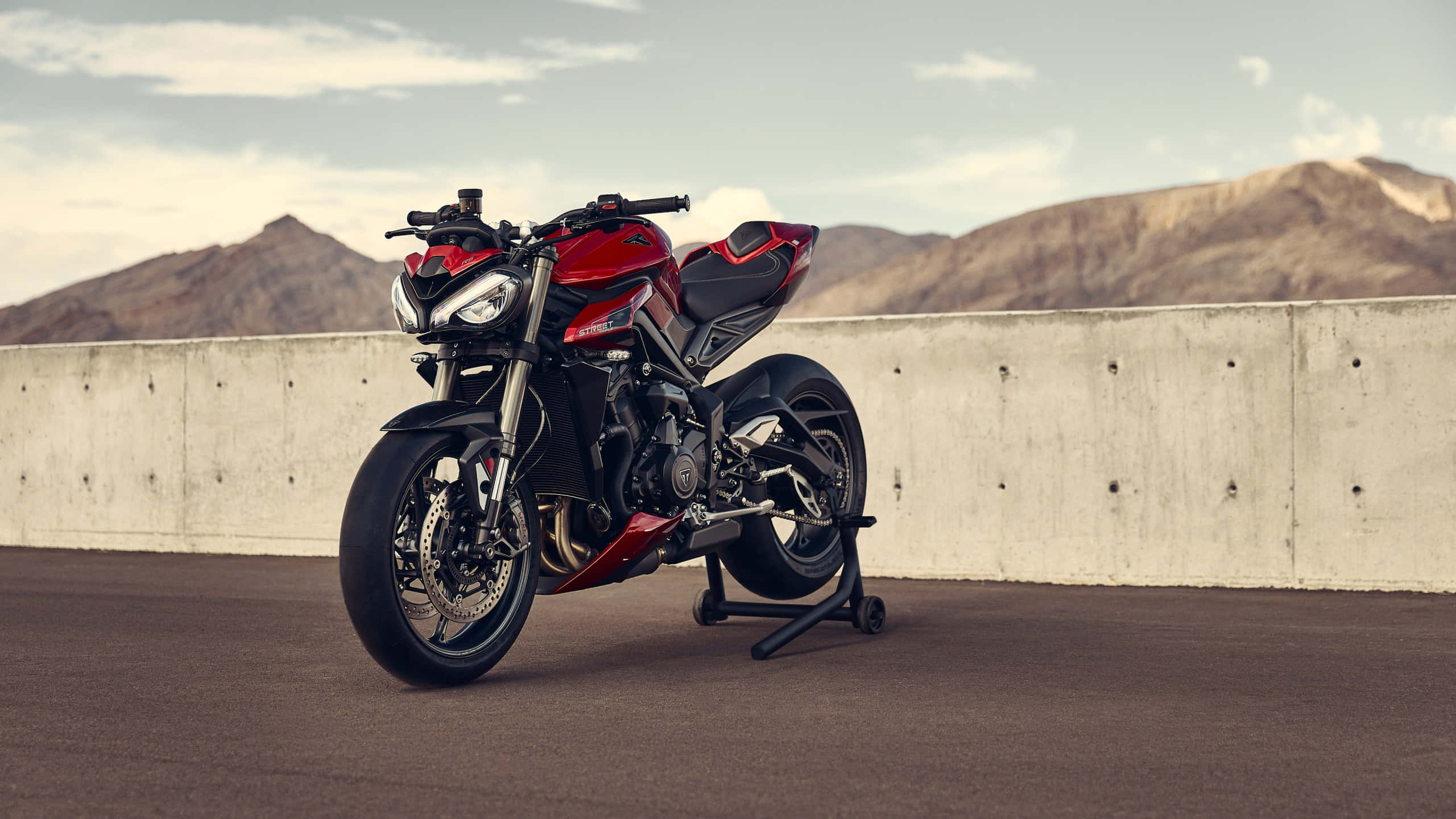Triumph Street Triple R機車背景 壁紙 — 一架時尚的紅色電單車停放在支架上，靠著混凝土牆，背景是崎嶇的山脈，天空多雲。