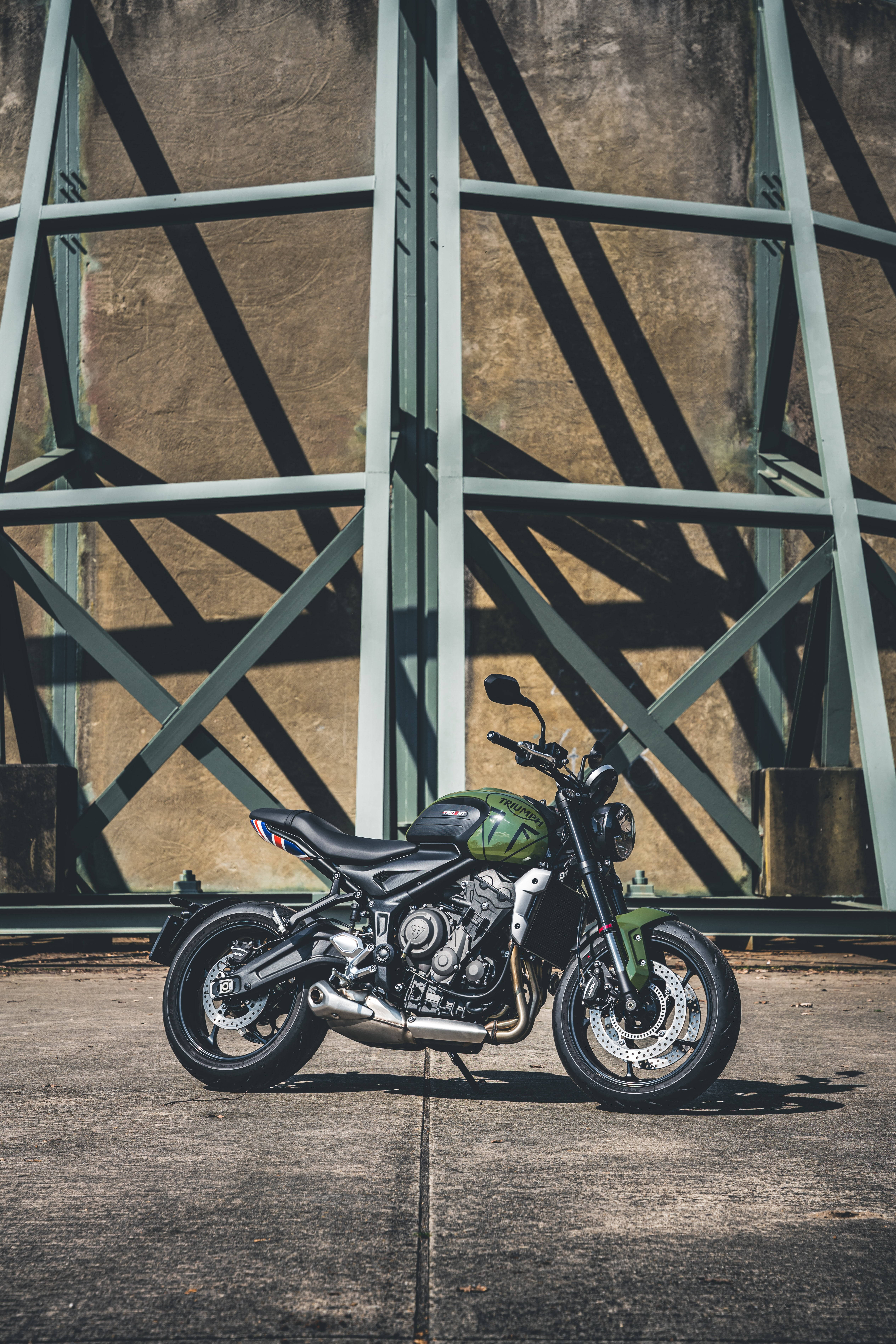 Triumph Trident 900 Bobber摩托車 壁紙 — 一架啞光綠色電單車喺幾何鋼樑下擺好姿勢，鋼樑喺混凝土上投下銳利嘅陰影。