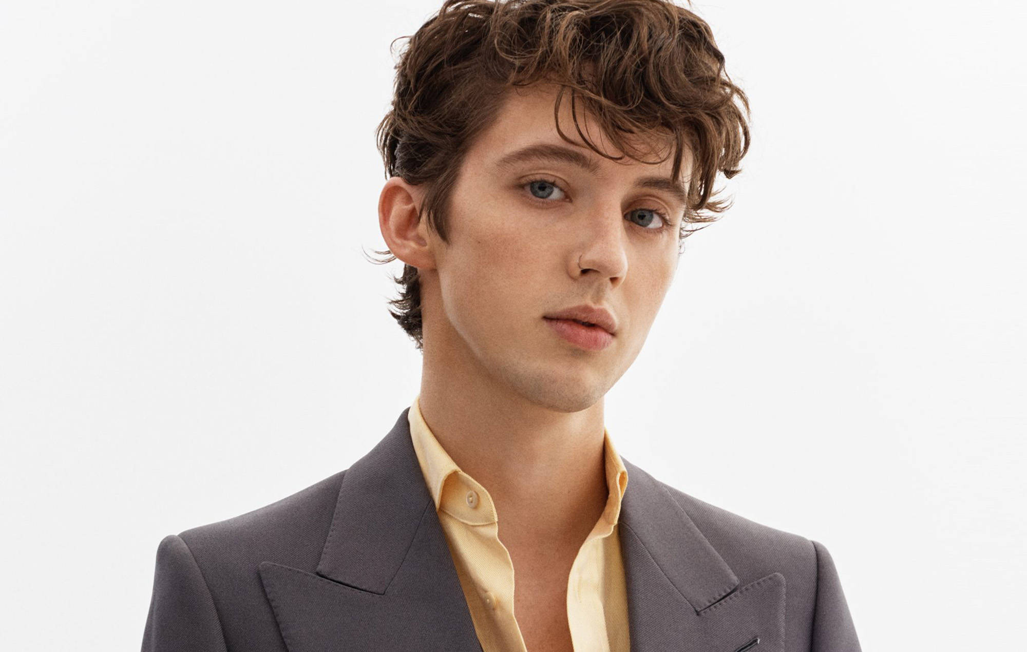 Troye Sivan Billboard 音樂獎肖像 壁紙 — 一位擁有棕色捲髮和藍色眼睛的年輕男士，在純白色背景前穿著灰色西裝外套和黃色襯衫。