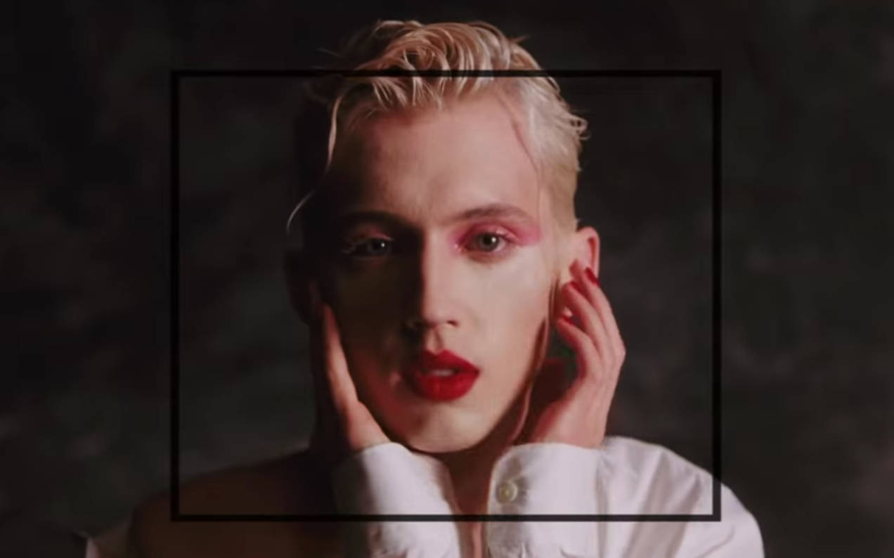 Troye Sivan《Bloom》MV 美貌 壁紙 — 一位擁有鉑金色頭髮和鮮豔紅唇的人，在深色紋理背景前用手框住臉龐，凝視著鏡頭。