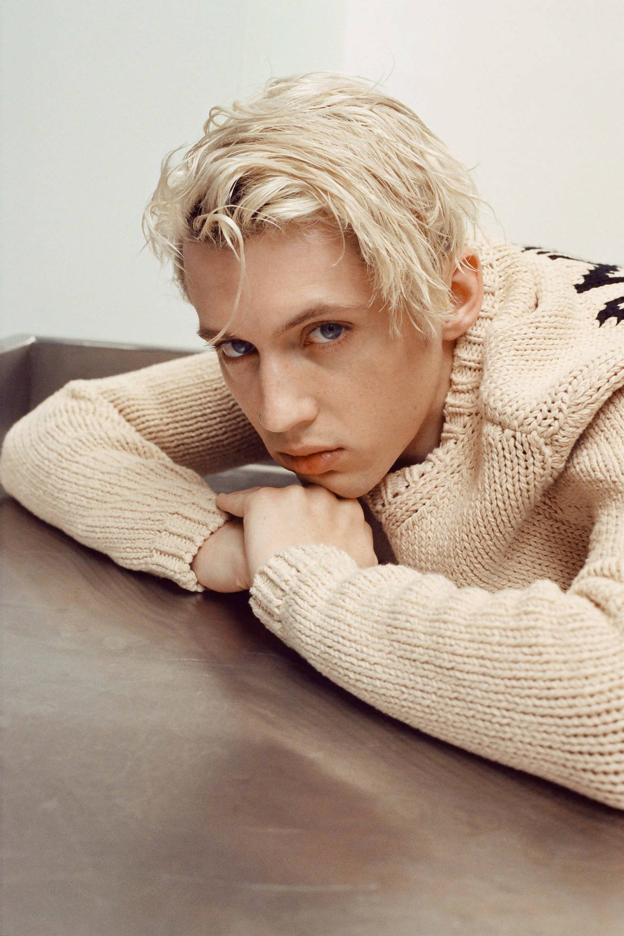 Troye Sivan 金髮帥氣造型 壁紙 — 一位年輕男子留著鉑金色頭髮，身穿米色針織毛衣，將下巴靠在手上。