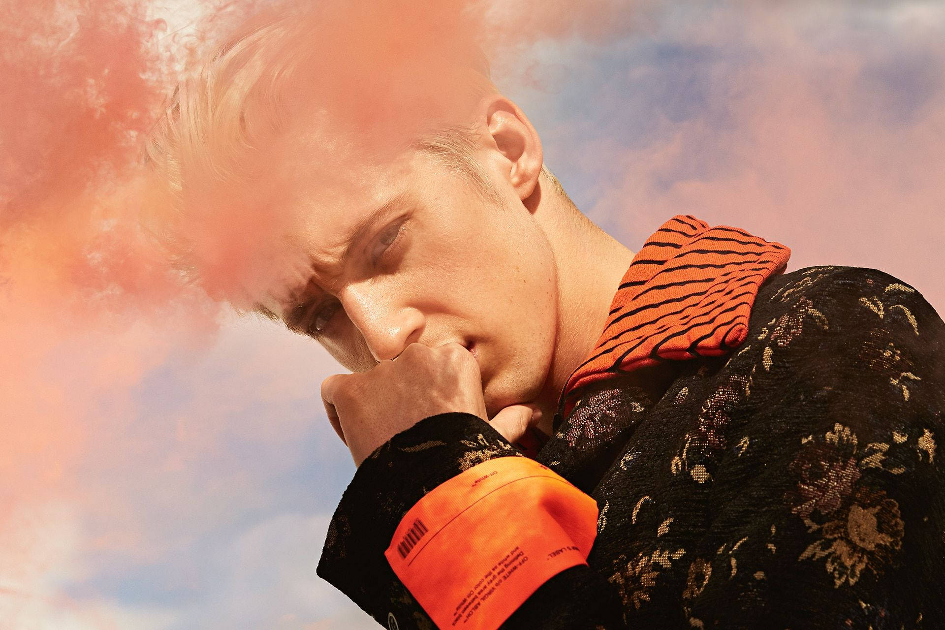 Troye Sivan GQ 雜誌拍攝 壁紙 — 一位擁有鉑金色頭髮和銳利眼神的年輕男子，穿著一件黑色花卉圖案外套，搭配橙色虎紋領，背景是夢幻的粉藍色天空。