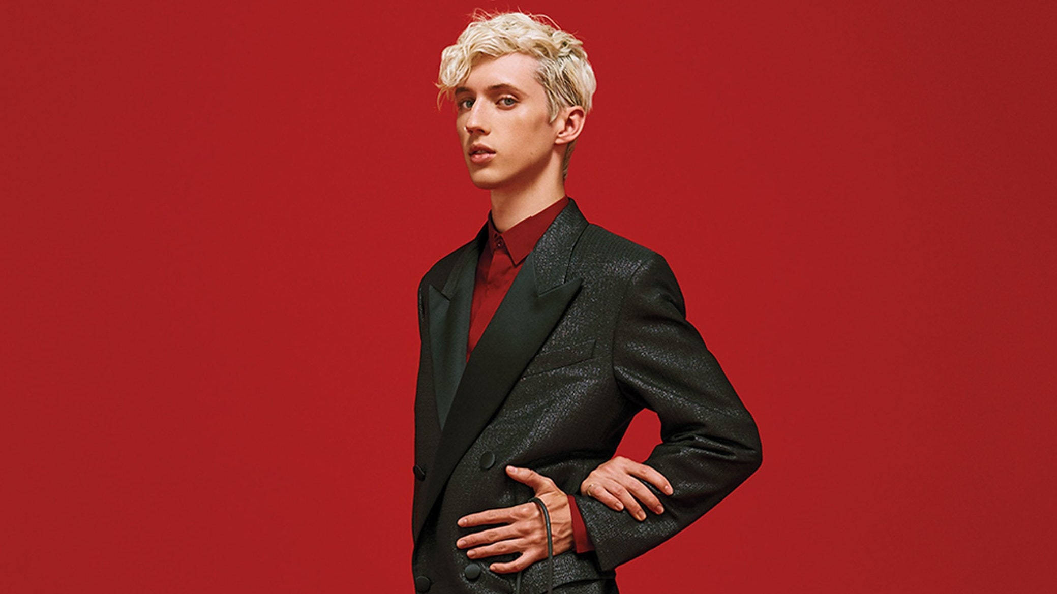 Troye Sivan 熱辣紅色造型 壁紙 — 一位時尚的年輕男子，一頭鉑金色頭髮，穿著質感黑色西裝外套，在鮮紅色的工作室背景前自信地擺姿勢。