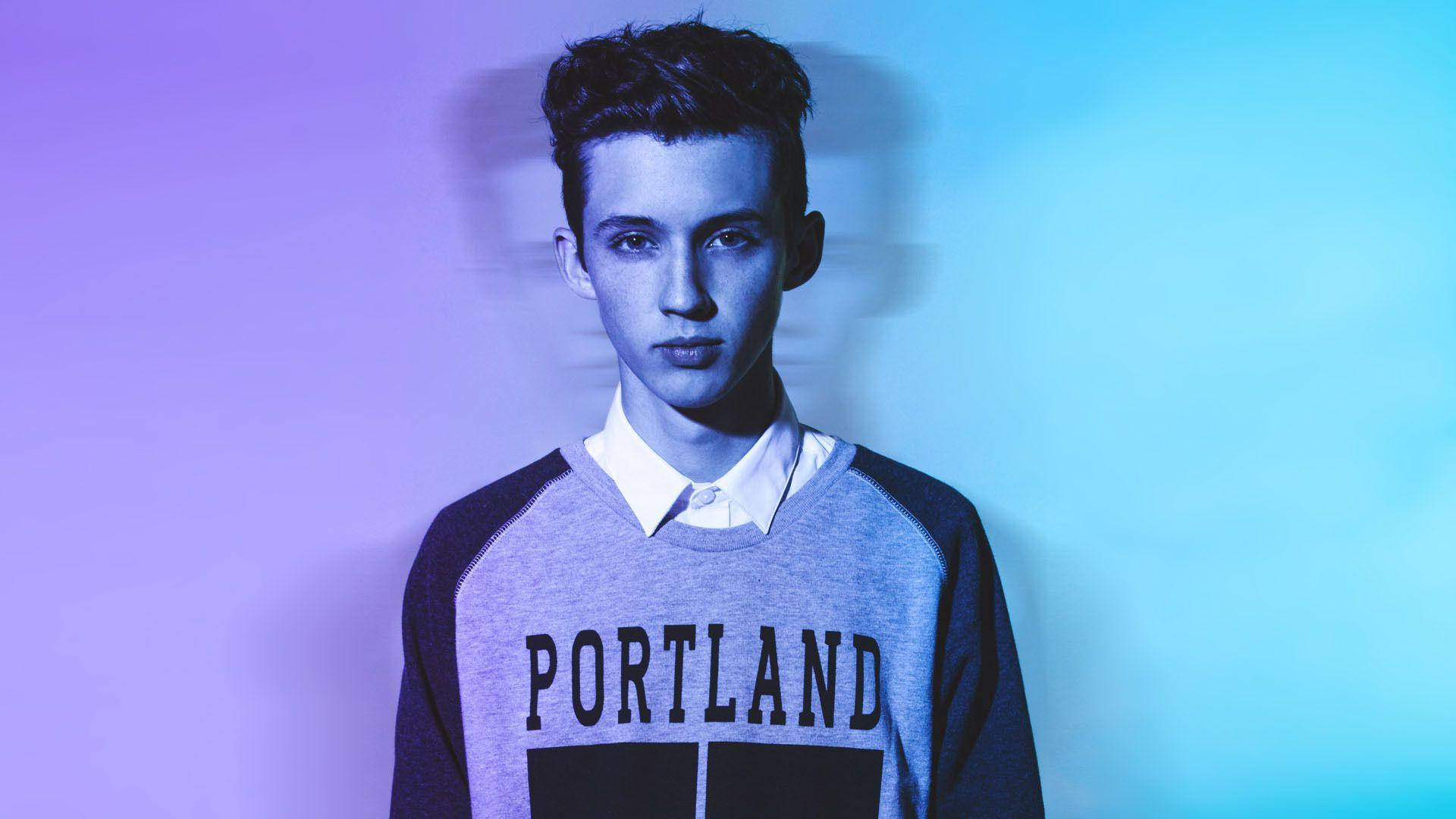 Troye Sivan 年輕藝術家 壁紙 — 一位穿著波特蘭衛衣的年輕男子站在柔和燈光的紫藍漸變背景前。
