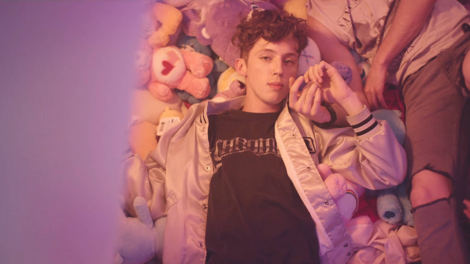 Troye Sivan《Youth》MV 場景 壁紙 — 一位捲髮年輕男子躺在紫色燈光下，周圍環繞著色彩繽紛的毛絨動物。