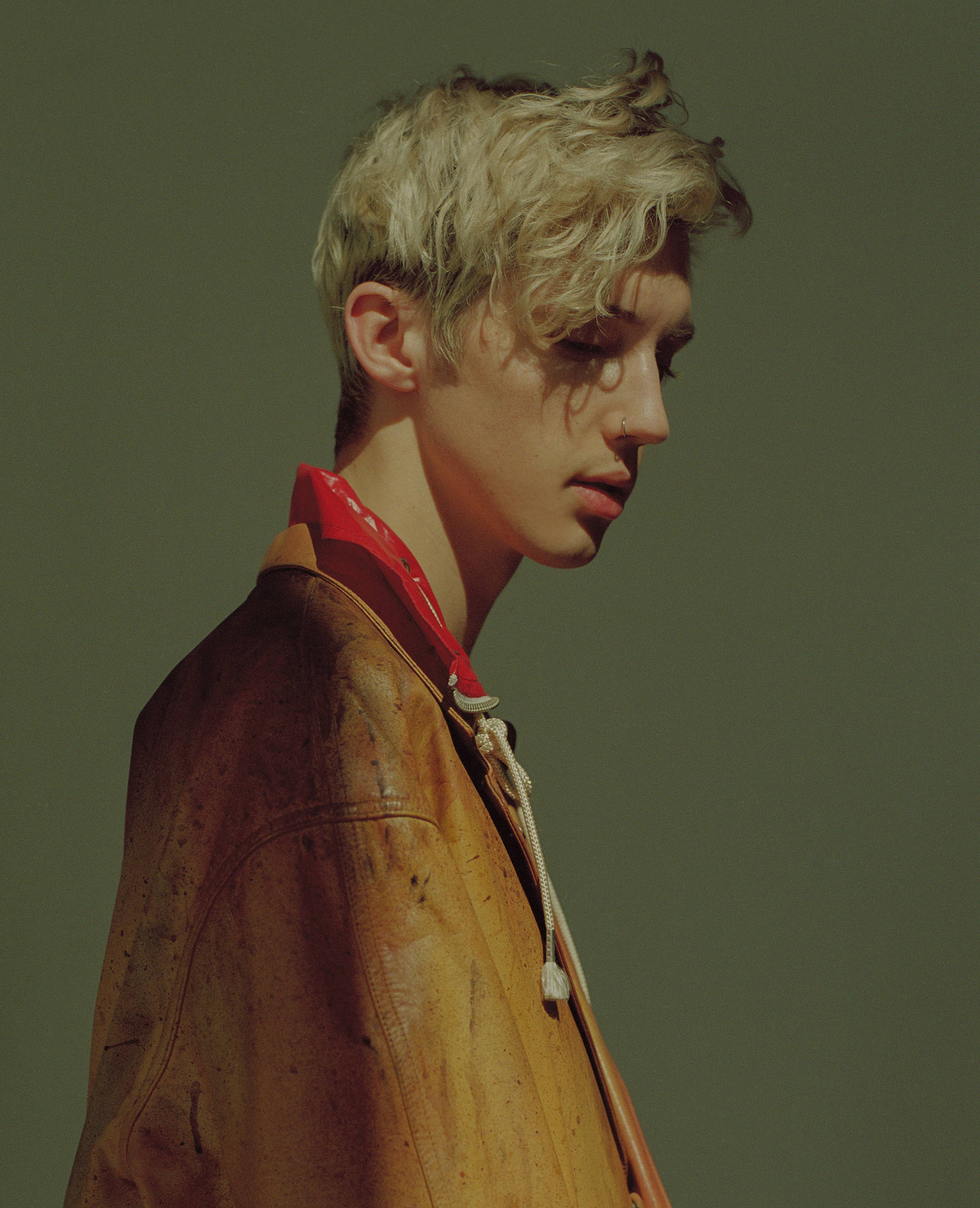 Troye Sivan 复古風情 壁紙 — 一位年輕男子，一頭凌亂的金髮，身穿一件破舊的棕色皮褸，內搭紅色襯衫，背景是柔和的綠色。
