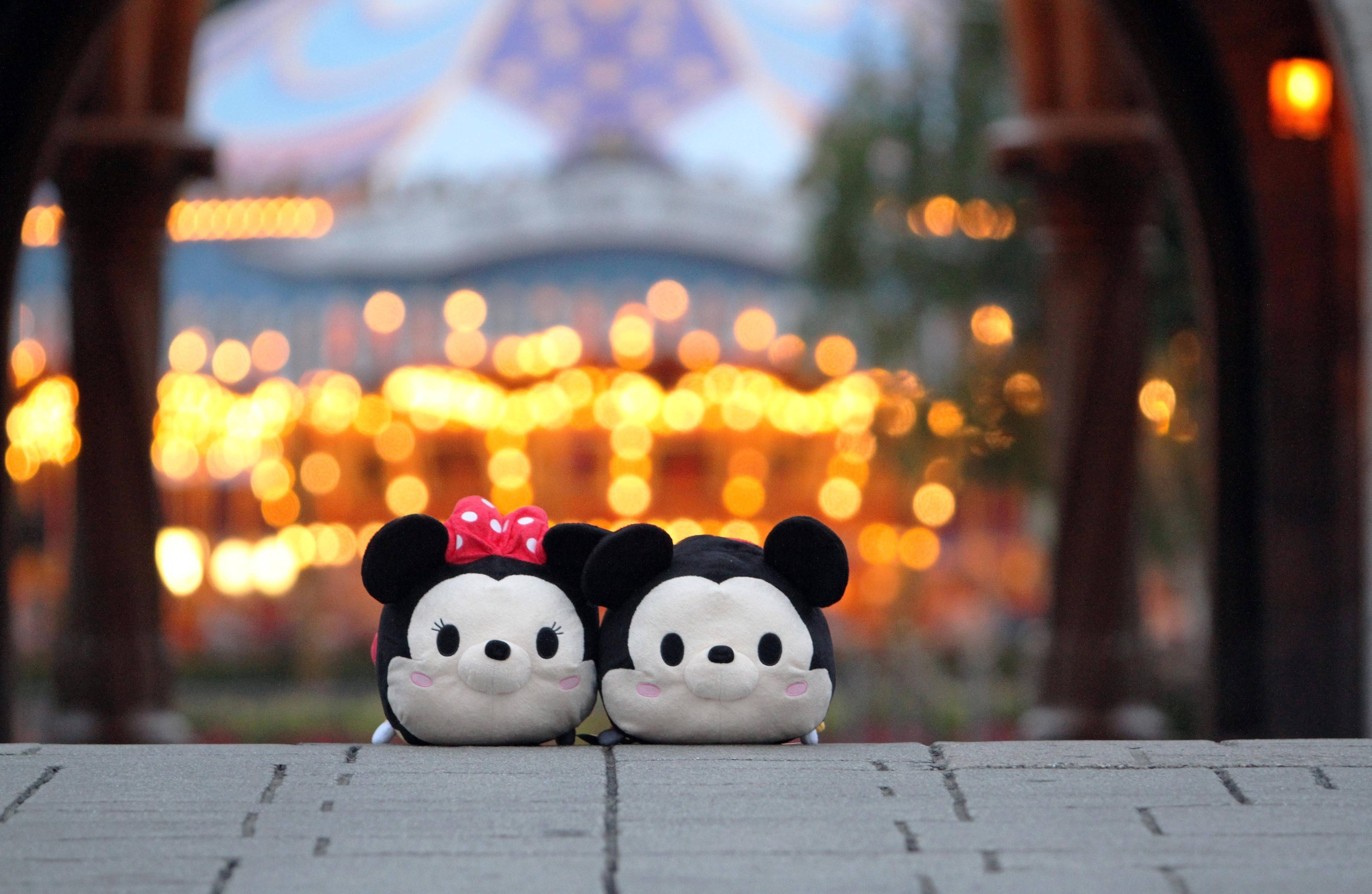 Tsum Tsum 遊樂場 壁紙 — 兩個可愛的毛絨米妮和米奇老鼠並排坐在石台上，背後是溫暖燈光下的遊樂園，呈現柔和散景效果。