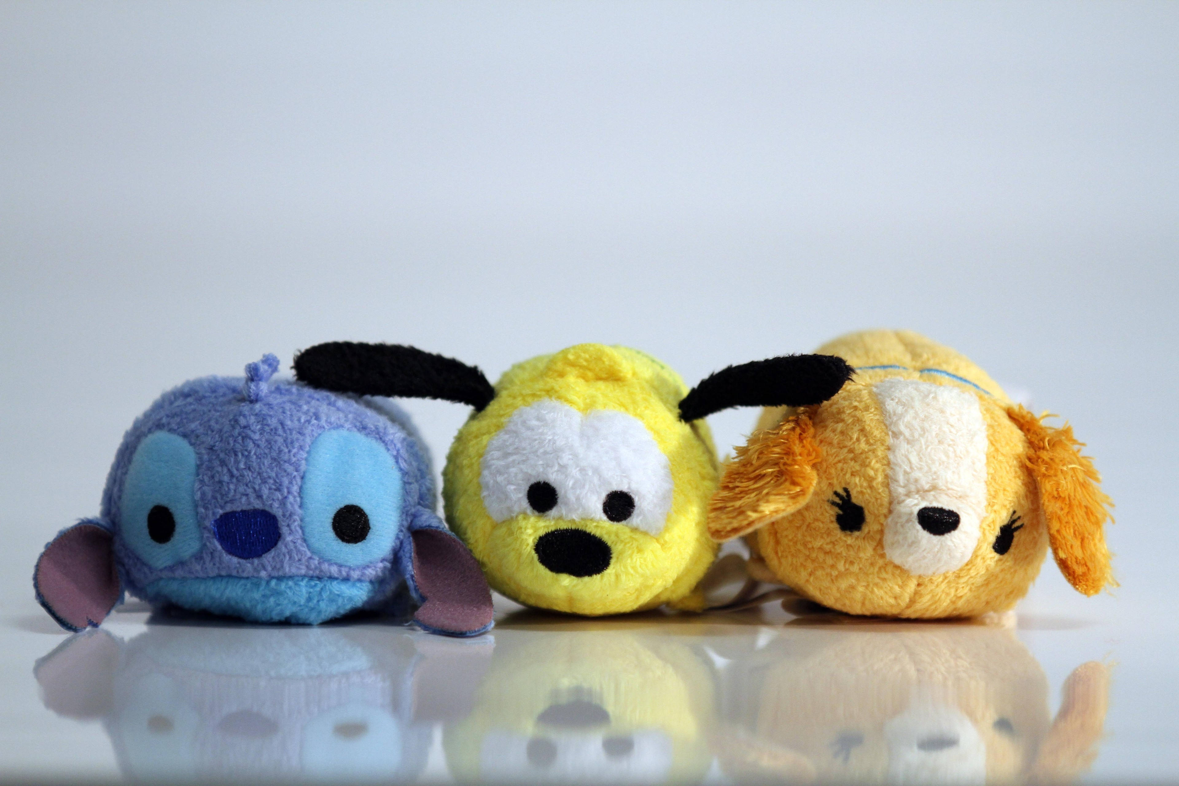 Tsum Tsum 狗狗 壁紙 — 三個毛絨迪士尼Tsum Tsum角色，藍色、黃色和橙色，並排坐在反光的白色表面上。