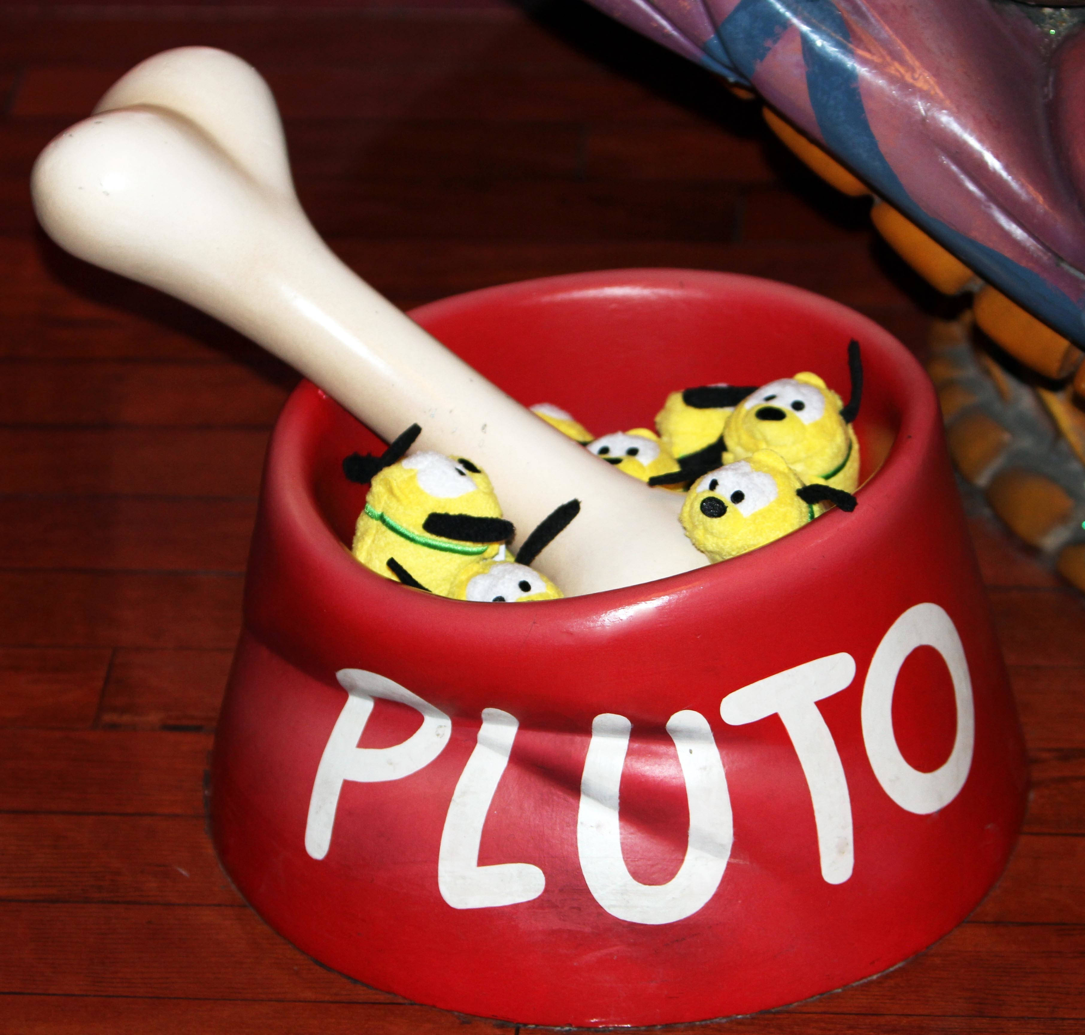 Tsum Tsum 普魯托 壁紙 — 一個紅色狗碗標有Pluto，內有黃色毛絨玩具和白色骨頭，放在木地板上。