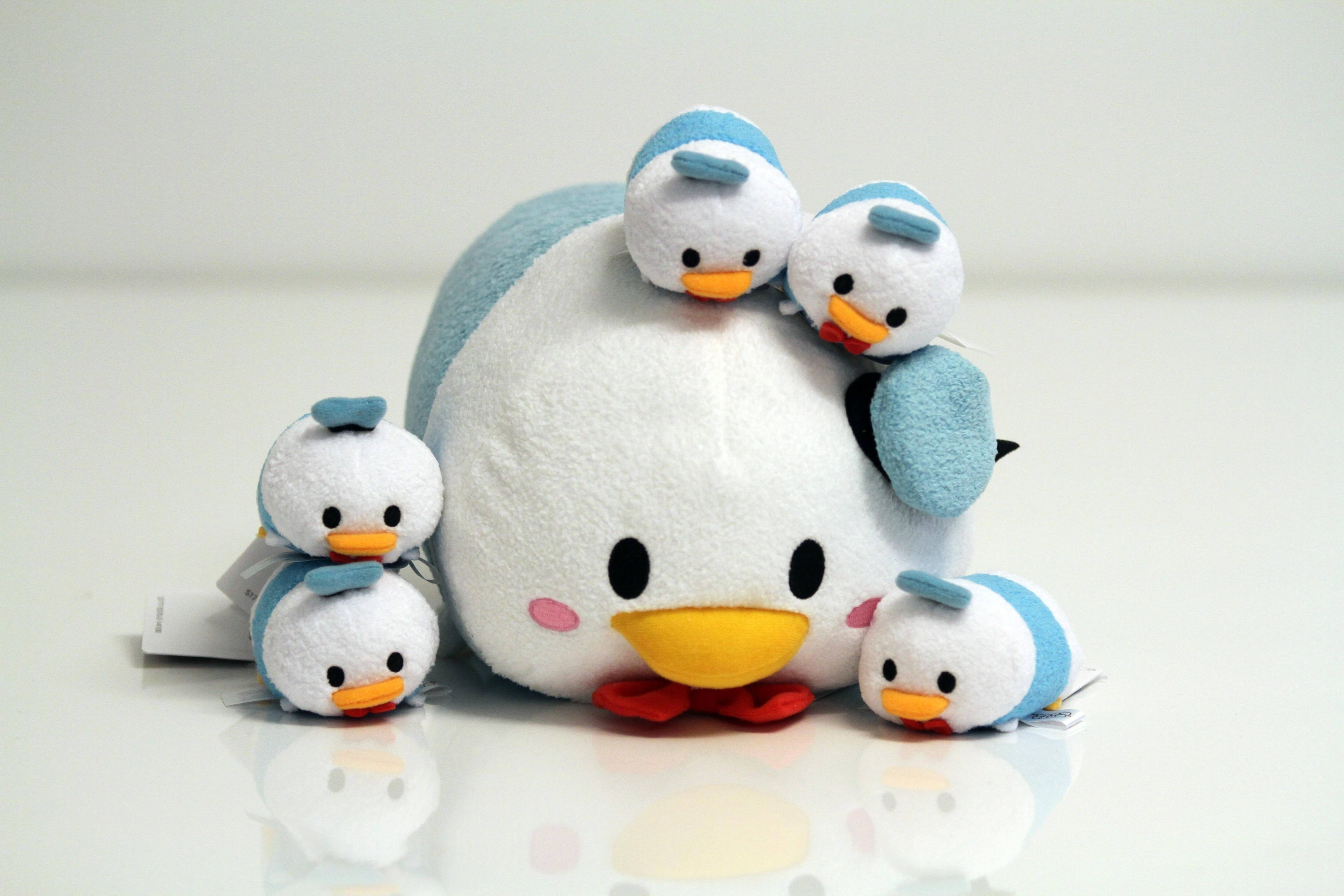 Tsum Tsum 唐老鴨 壁紙 — 一隻戴藍色帽子和紅色蝴蝶結的大型白色鴨子毛絨玩具，被五個較小的相同鴨子毛絨玩具包圍，放在反光的白色表面上。