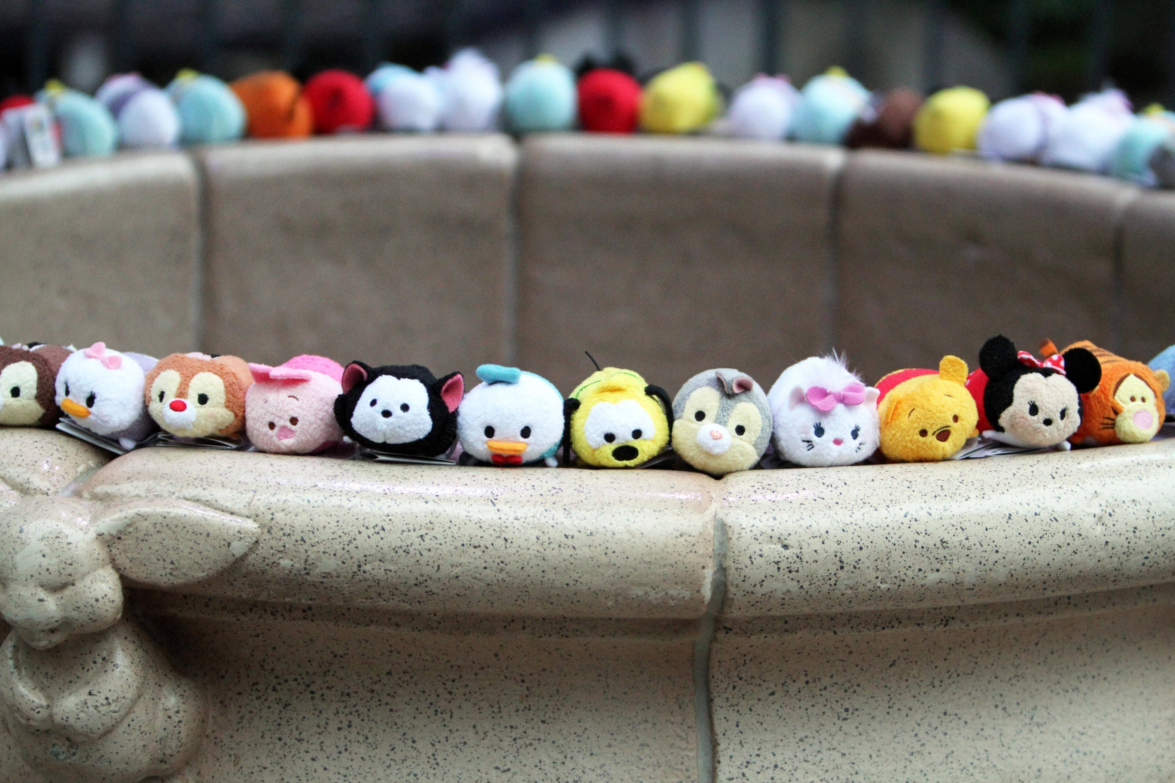 Tsum Tsum 噴泉 壁紙 — 一排色彩繽紛的迪士尼 Tsum Tsum 毛絨公仔整齊地坐在斑點石噴泉邊緣上。