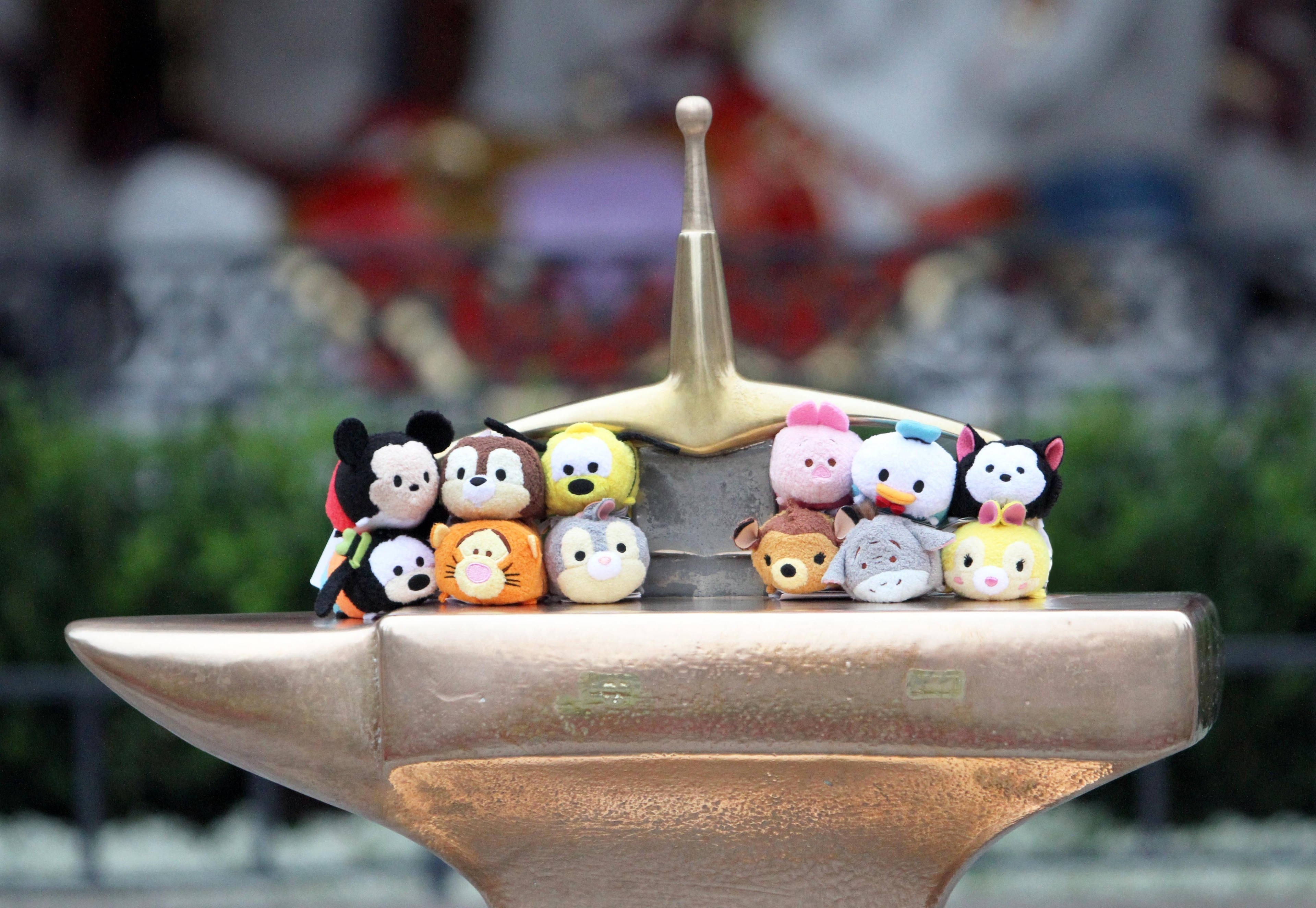 Tsum Tsum 展示 壁紙 — 一組色彩繽紛的迪士尼 Tsum Tsum 毛絨公仔坐在金色鐵砧上，背景是模糊的公園。