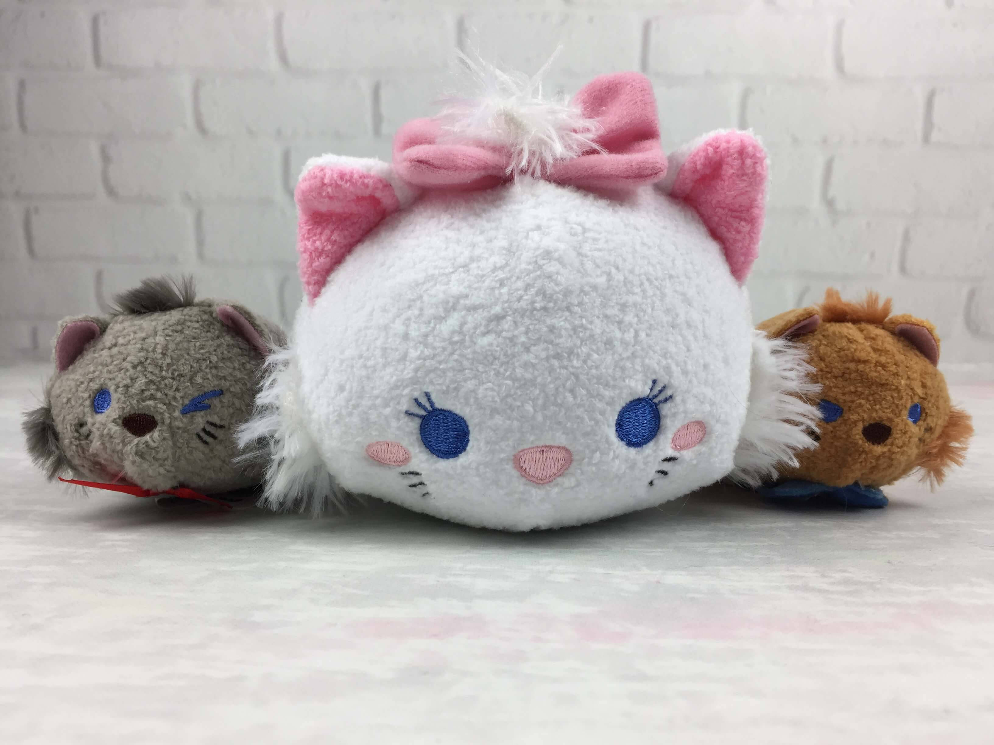 Tsum Tsum 王子貓 壁紙 — 三個可愛的Tsum Tsum毛絨玩具，包括一隻戴粉紅色蝴蝶結的白貓，坐在淺色表面上，背景是白色磚牆。